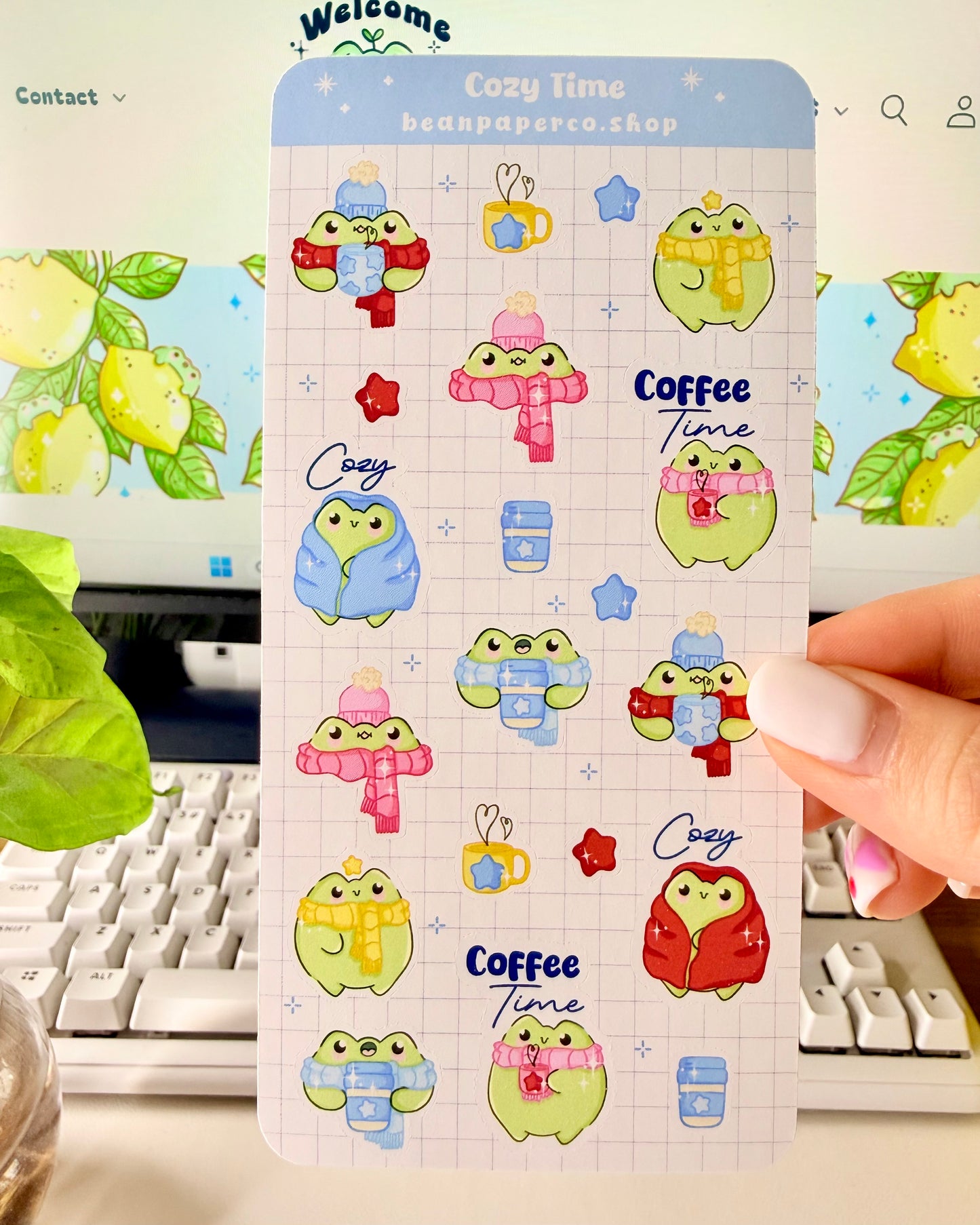 Hand holding a cozy frog sticker sheet featuring cute green frogs in scarves and hats, holding coffee cups, and wrapped in blankets.
