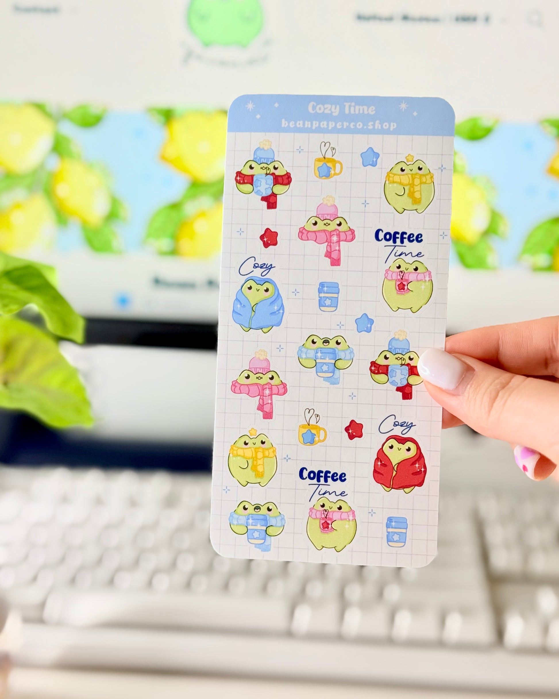 Hand holding a cozy frog sticker sheet featuring cute green frogs in scarves and hats, holding coffee cups, and wrapped in blankets.