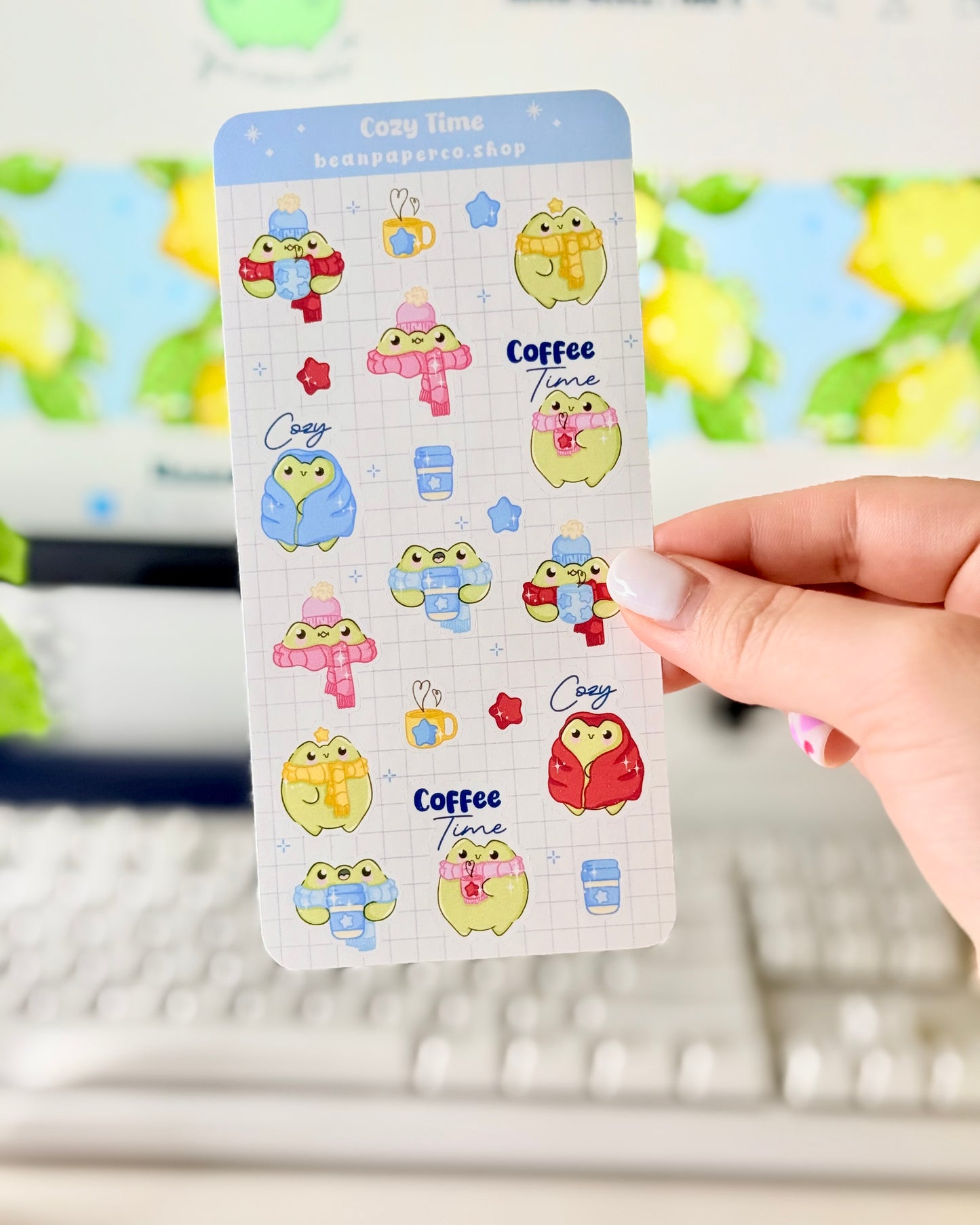 Hand holding a cozy frog sticker sheet featuring cute green frogs in scarves and hats, holding coffee cups, and wrapped in blankets.