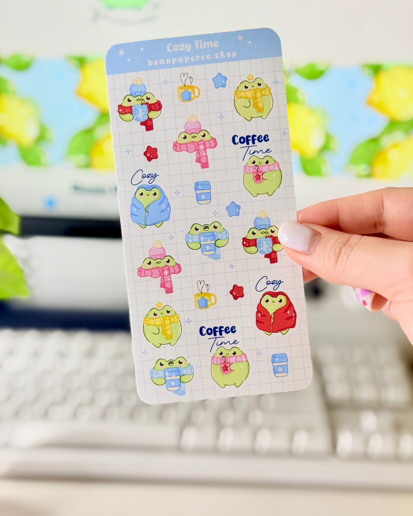 Hand holding a cozy frog sticker sheet featuring cute green frogs in scarves and hats, holding coffee cups, and wrapped in blankets.