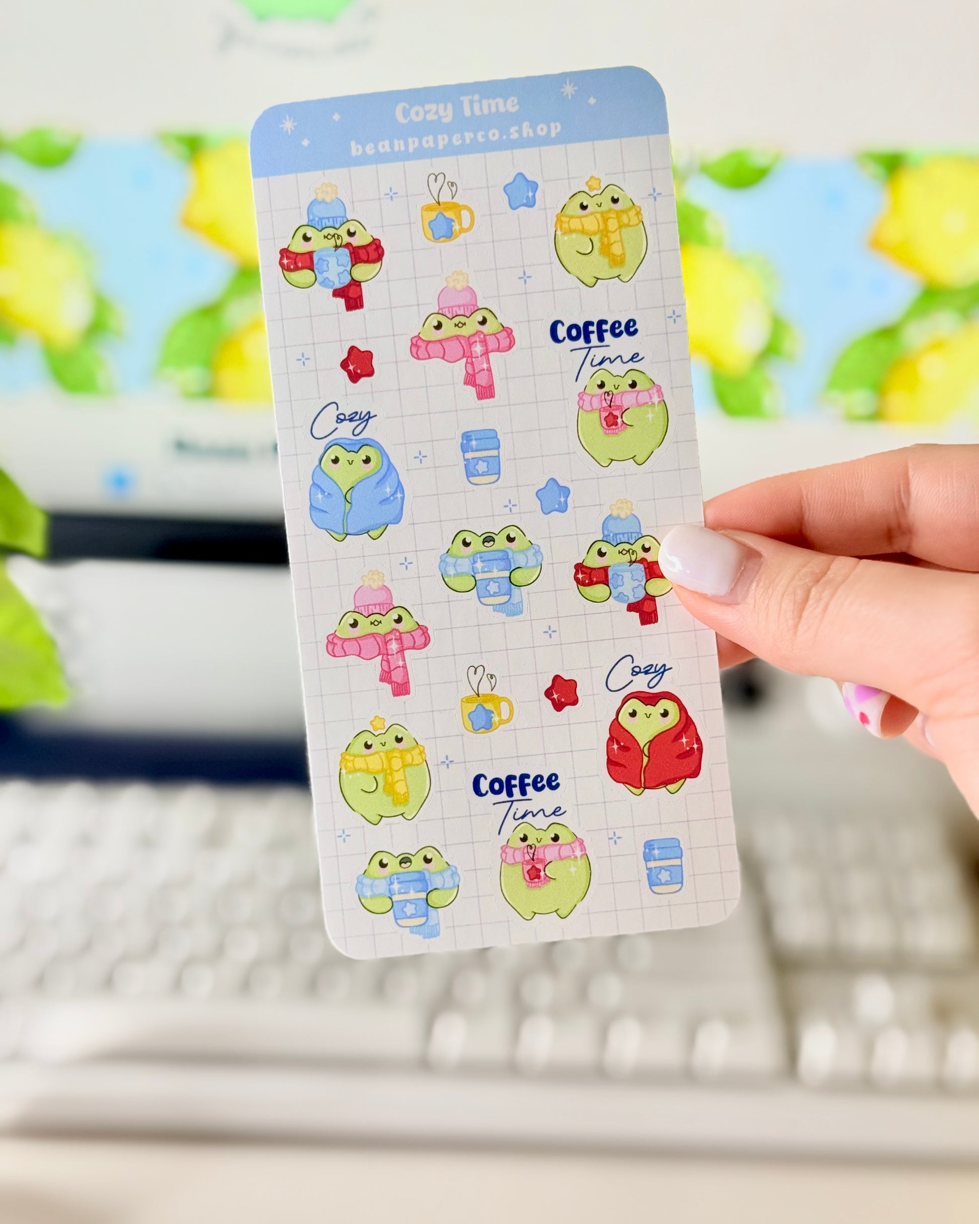 Hand holding a cozy frog sticker sheet featuring cute green frogs in scarves and hats, holding coffee cups, and wrapped in blankets.