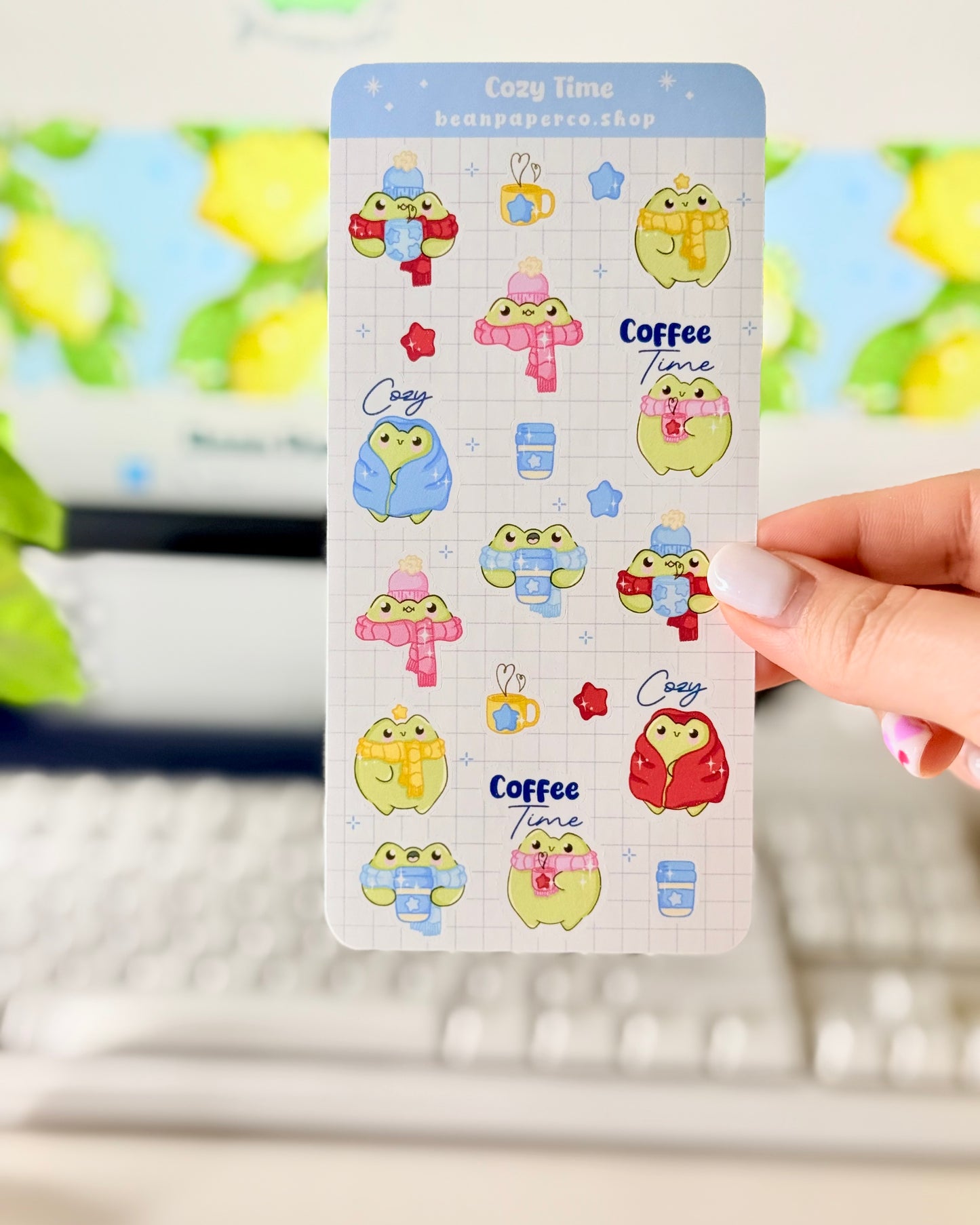 Hand holding a cozy frog sticker sheet featuring cute green frogs in scarves and hats, holding coffee cups, and wrapped in blankets.
