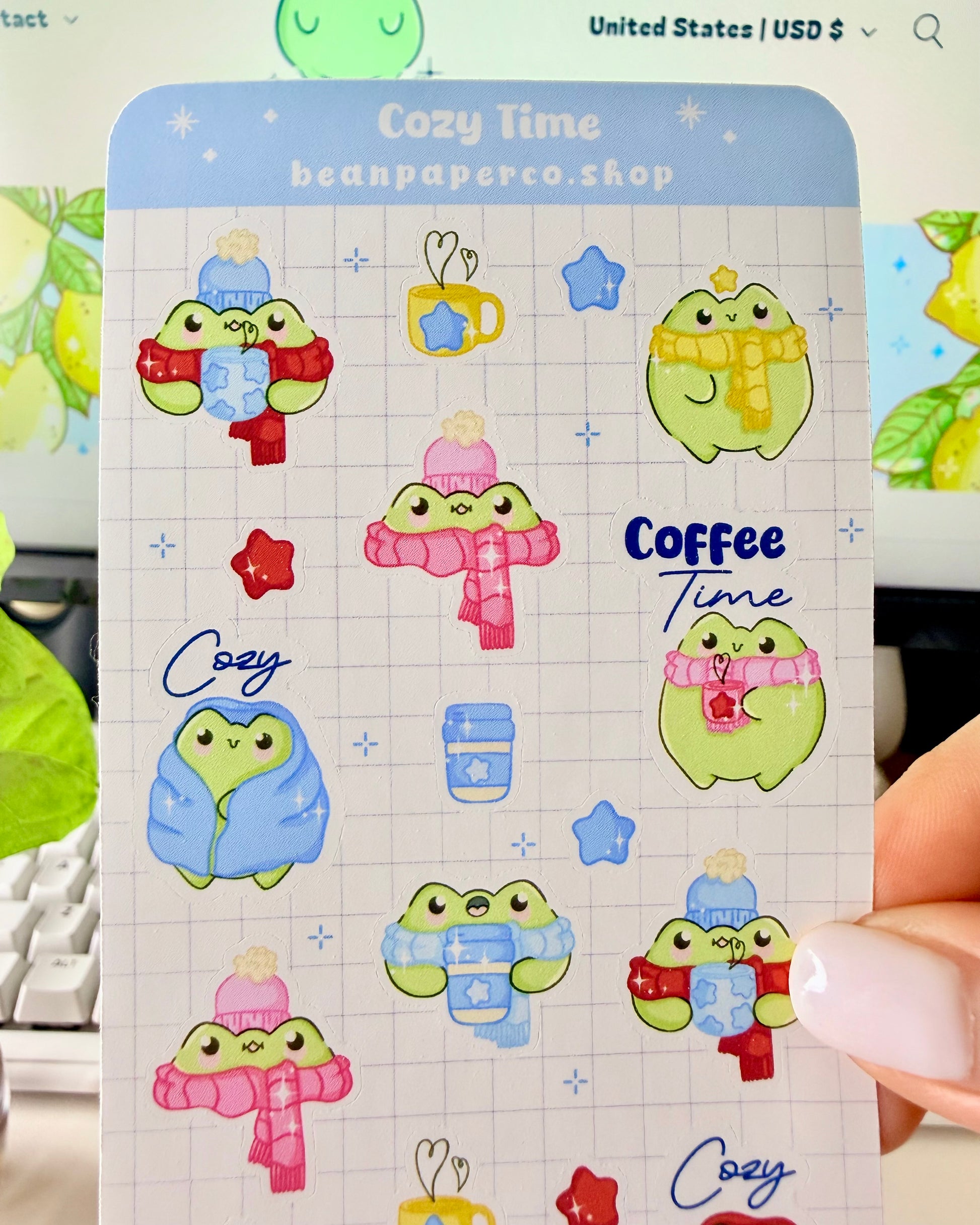 Hand holding a cozy frog sticker sheet featuring cute green frogs in scarves and hats, holding coffee cups, and wrapped in blankets.