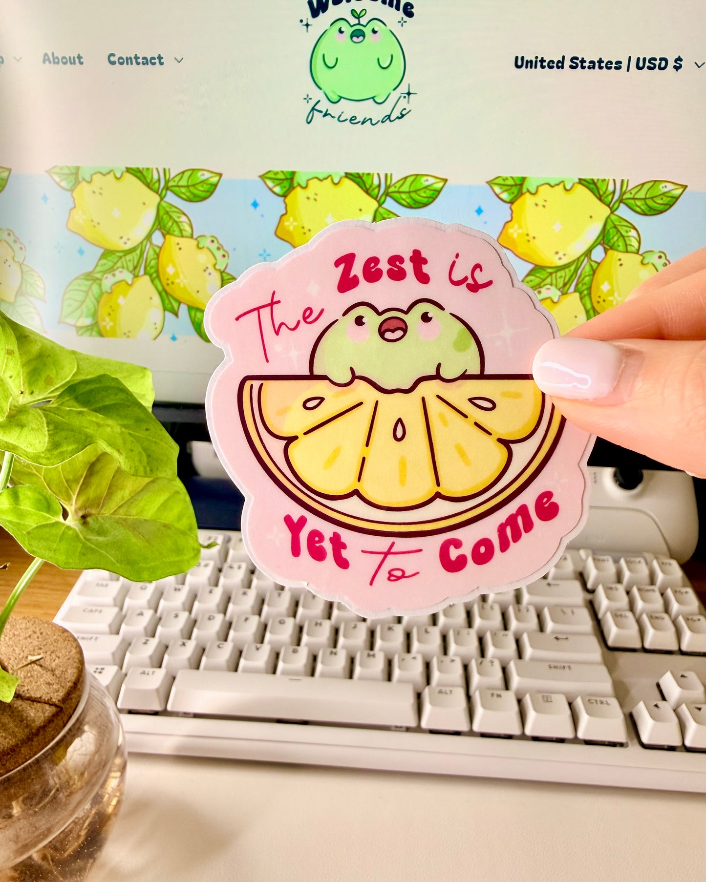 Kawaii Frog Lemon Vinyl Sticker with a cartoon frog character and text that says, "The Zest is Yet to Come," held by a hand in front of a computer screen.