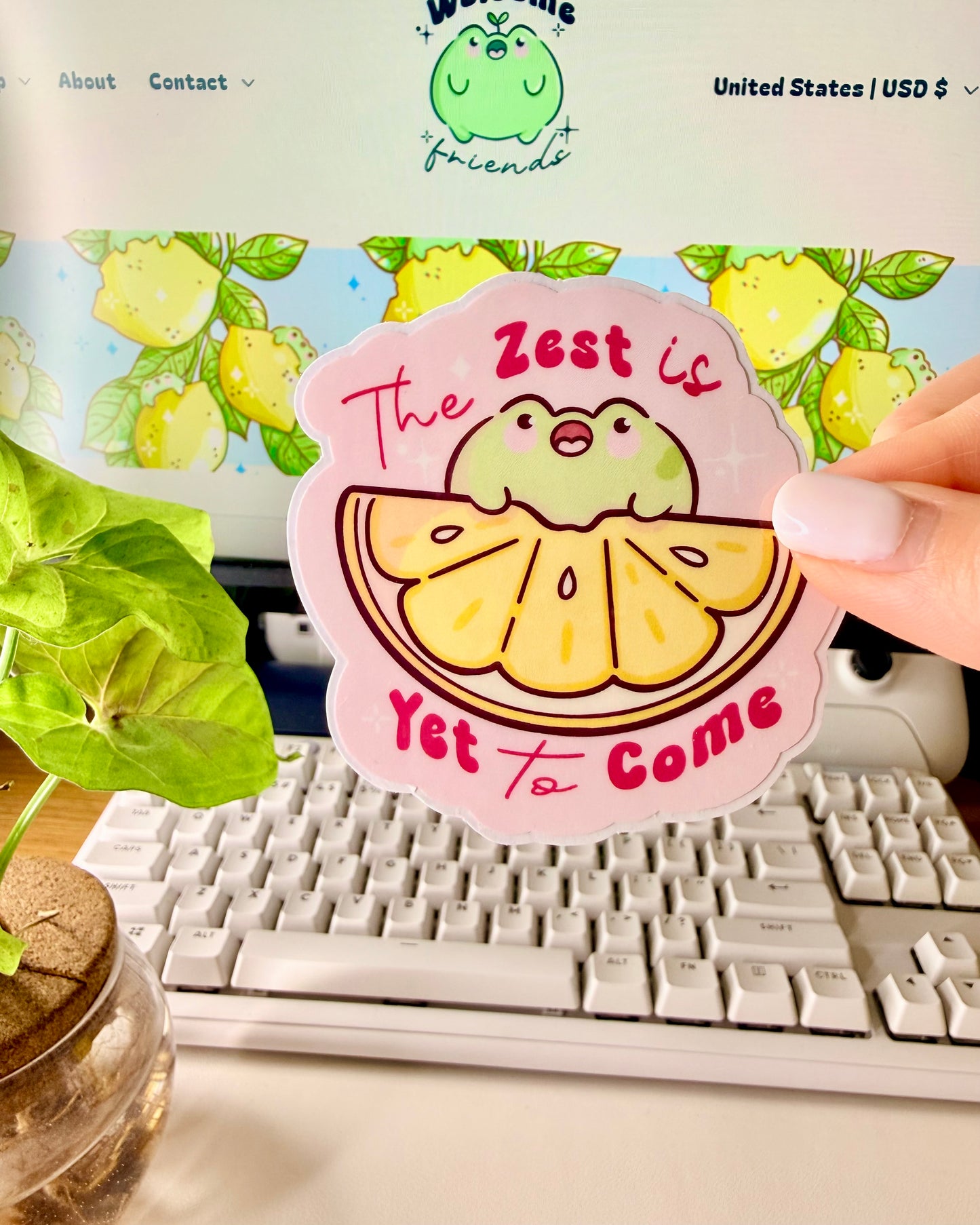 Kawaii Frog Lemon Vinyl Sticker with a cartoon frog character and text that says, "The Zest is Yet to Come," held by a hand in front of a computer screen.
