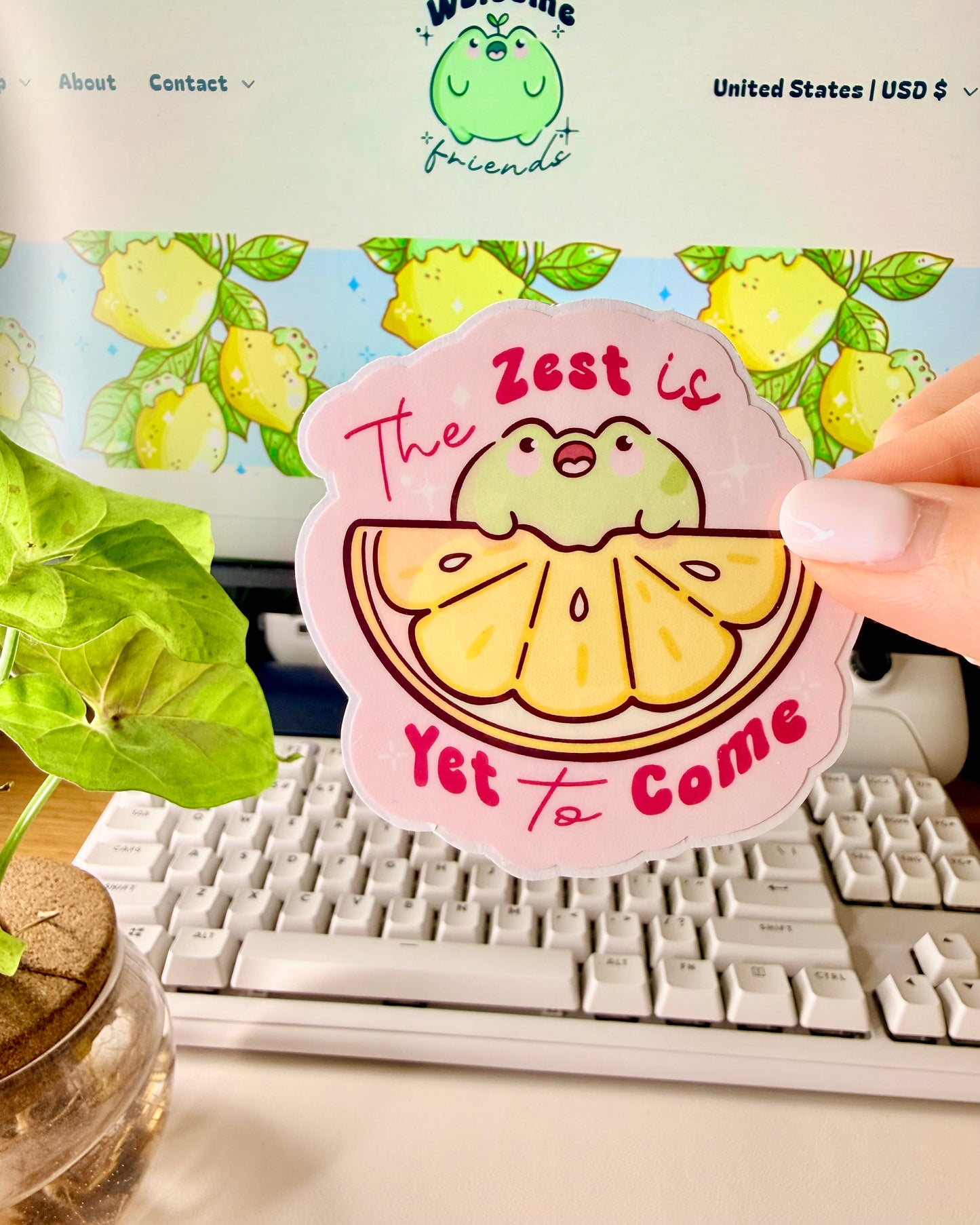 Kawaii Frog Lemon Vinyl Sticker with a cartoon frog character and text that says, "The Zest is Yet to Come," held by a hand in front of a computer screen.