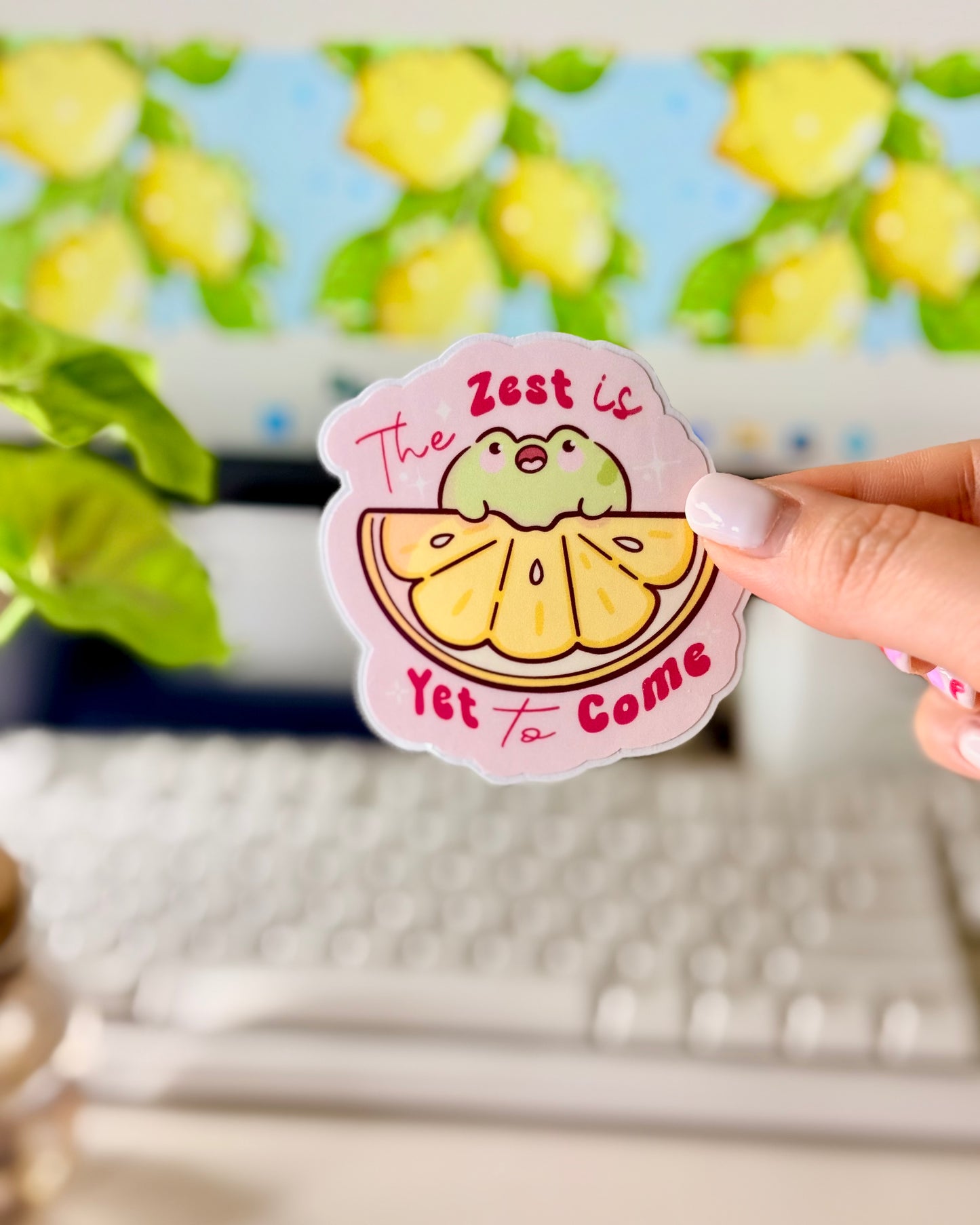 Kawaii Frog Lemon Vinyl Sticker with a cartoon frog character and text that says, "The Zest is Yet to Come," held by a hand in front of a computer screen.