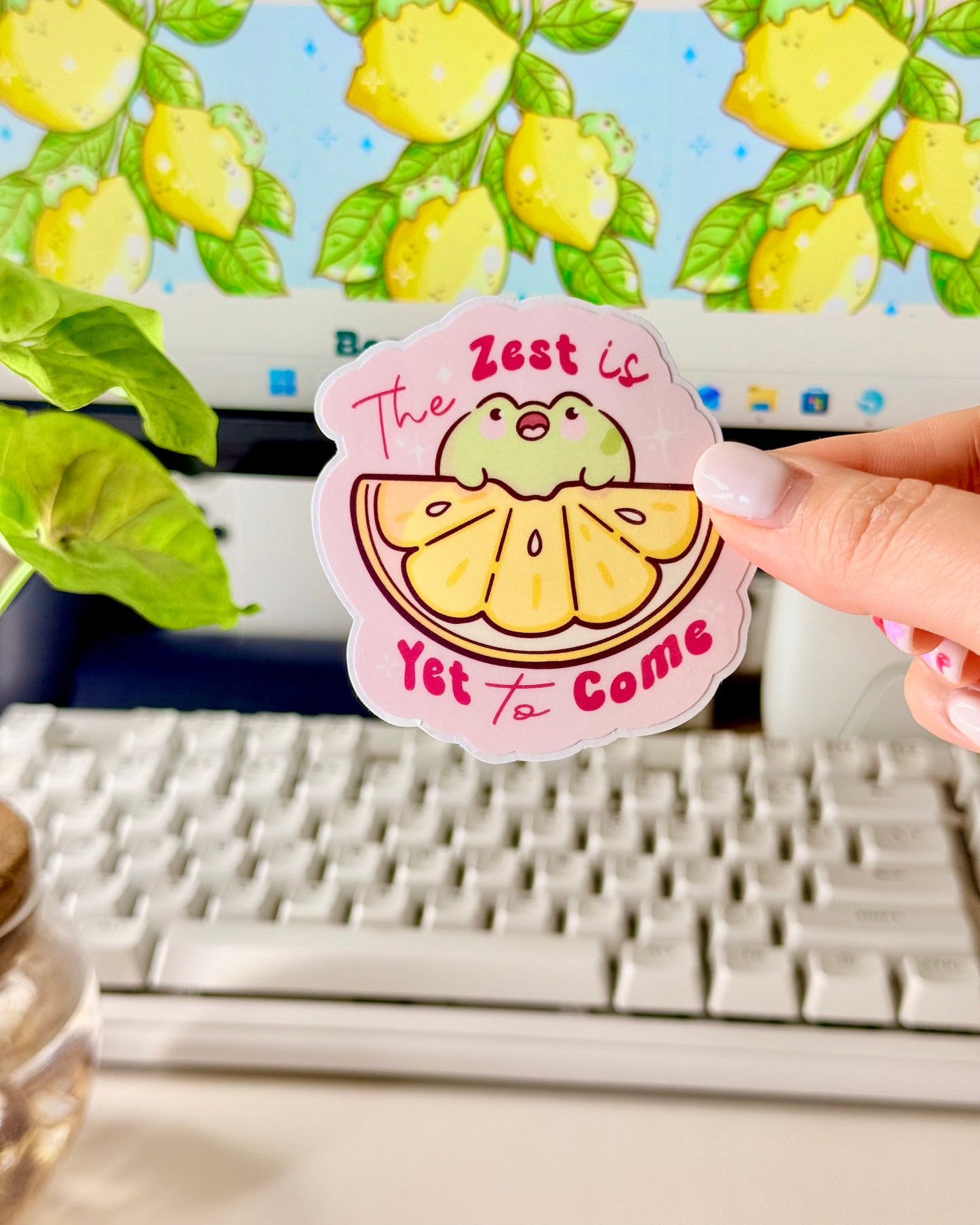 Kawaii Frog Lemon Vinyl Sticker with a cartoon frog character and text that says, "The Zest is Yet to Come," held by a hand in front of a computer screen.