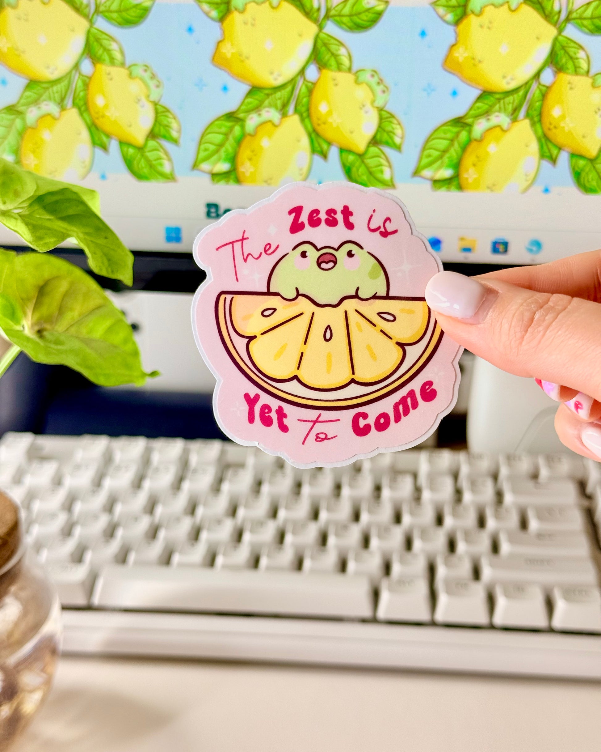 Kawaii Frog Lemon Vinyl Sticker with a cartoon frog character and text that says, "The Zest is Yet to Come," held by a hand in front of a computer screen.
