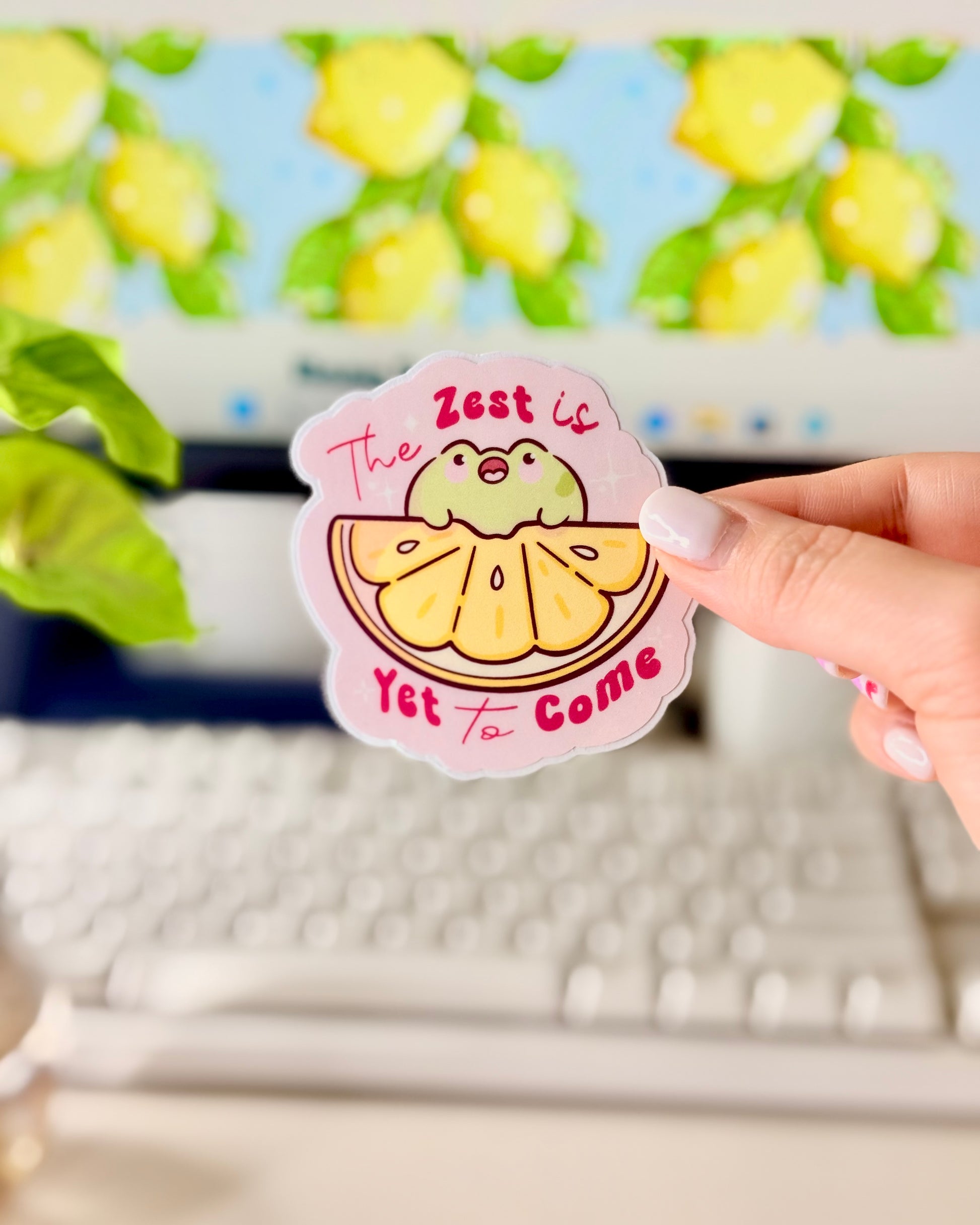 Kawaii Frog Lemon Vinyl Sticker with a cartoon frog character and text that says, "The Zest is Yet to Come," held by a hand in front of a computer screen.
