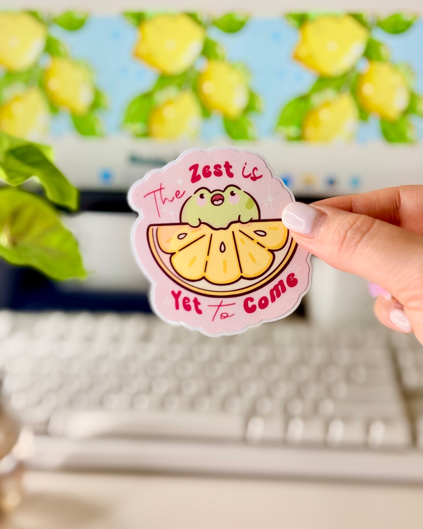 Kawaii Frog Lemon Vinyl Sticker with a cartoon frog character and text that says, "The Zest is Yet to Come," held by a hand in front of a computer screen.
