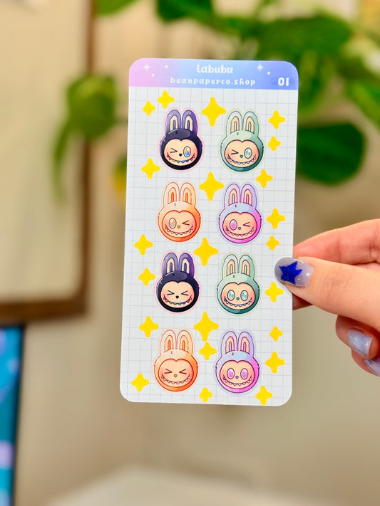 Labubu The Monster Sticker Sheet with black, blue green, orange, and purple labubus held by hand against a blurred background.