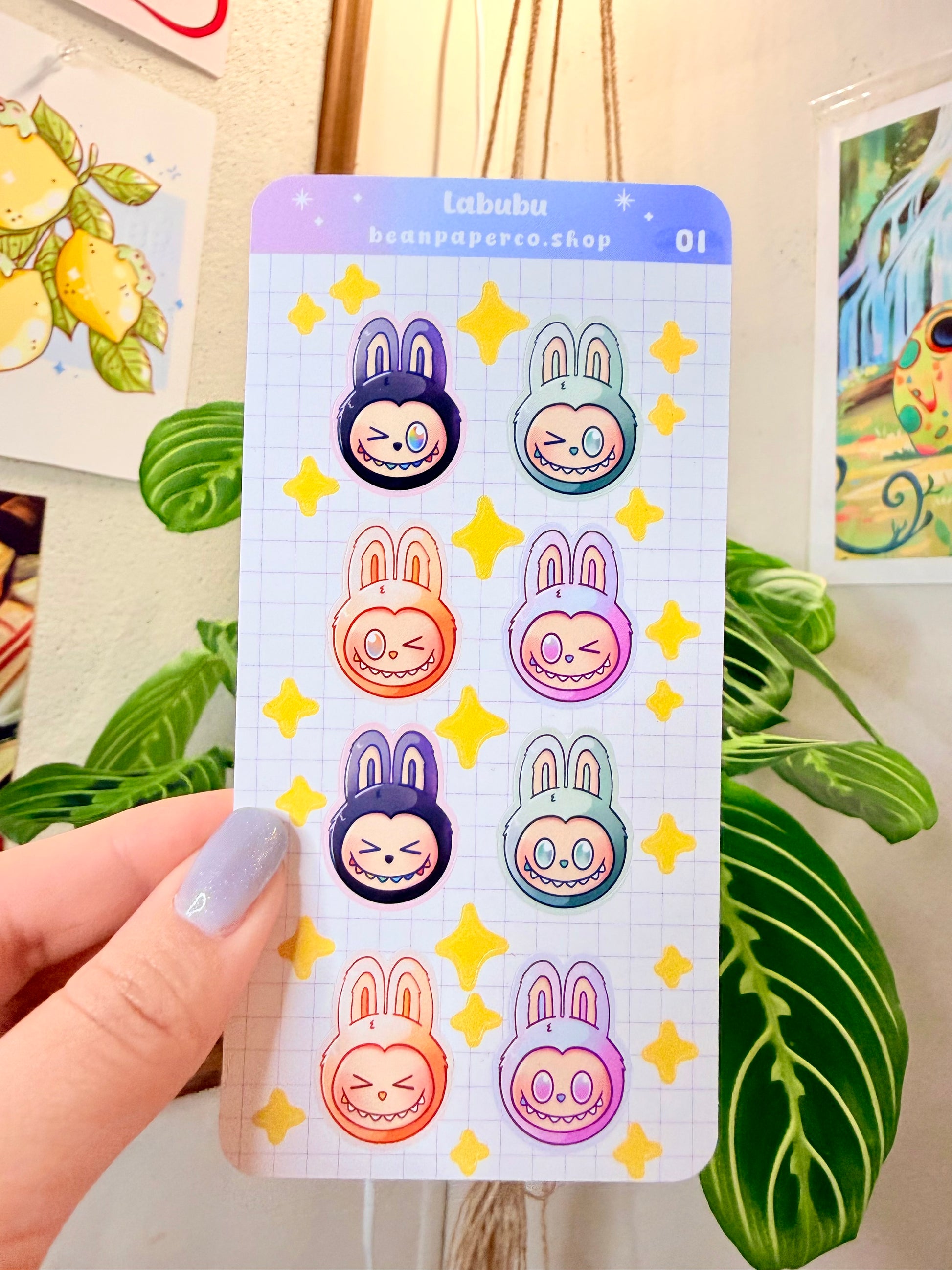 Labubu The Monster Sticker Sheet sheet with black, blue green, orange, and purple labubus held by hand with plant in the background.