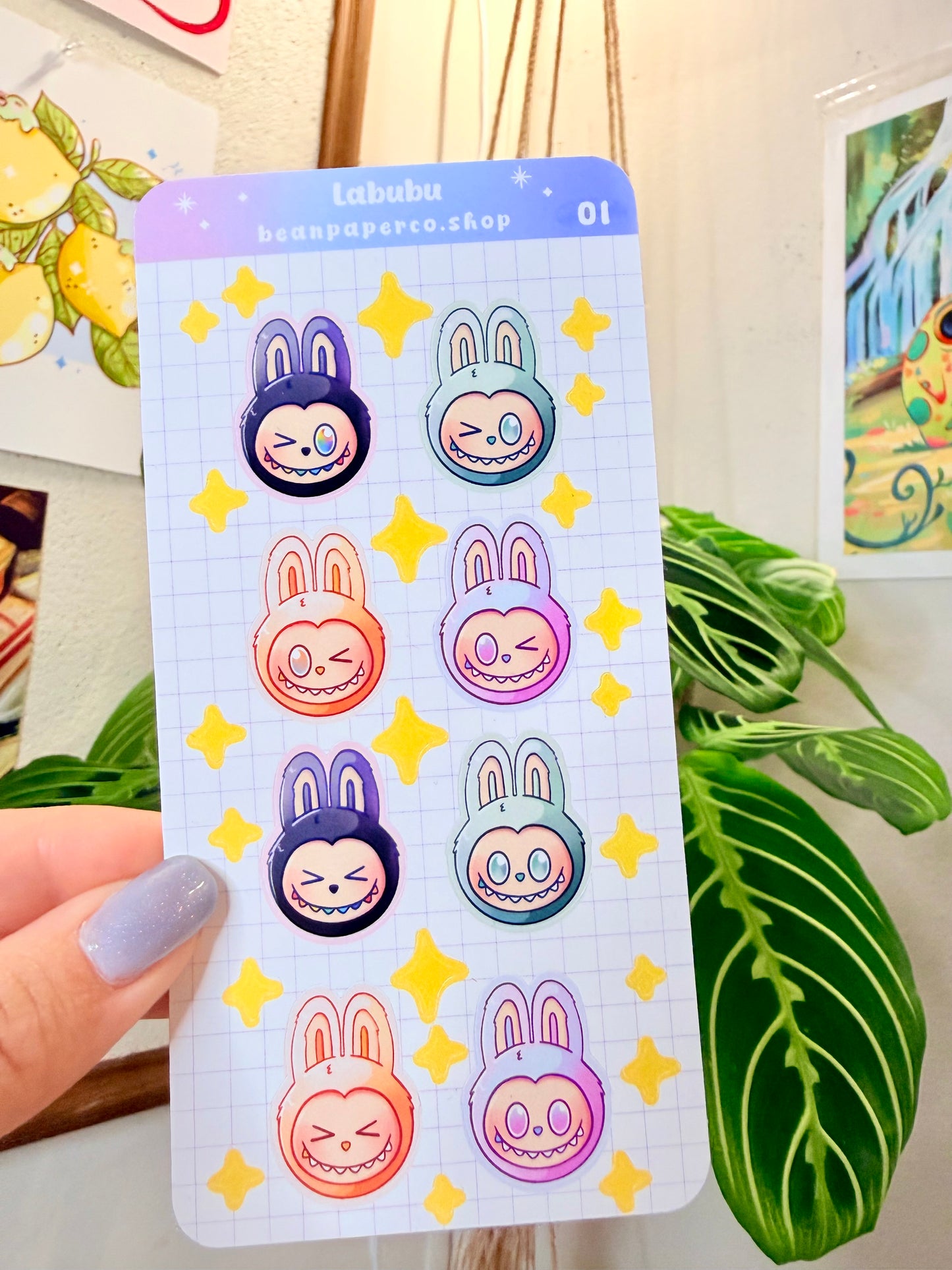 Labubu The Monster Sticker Sheet with black, blue green, orange, and purple labubus held by hand with plant in the background.