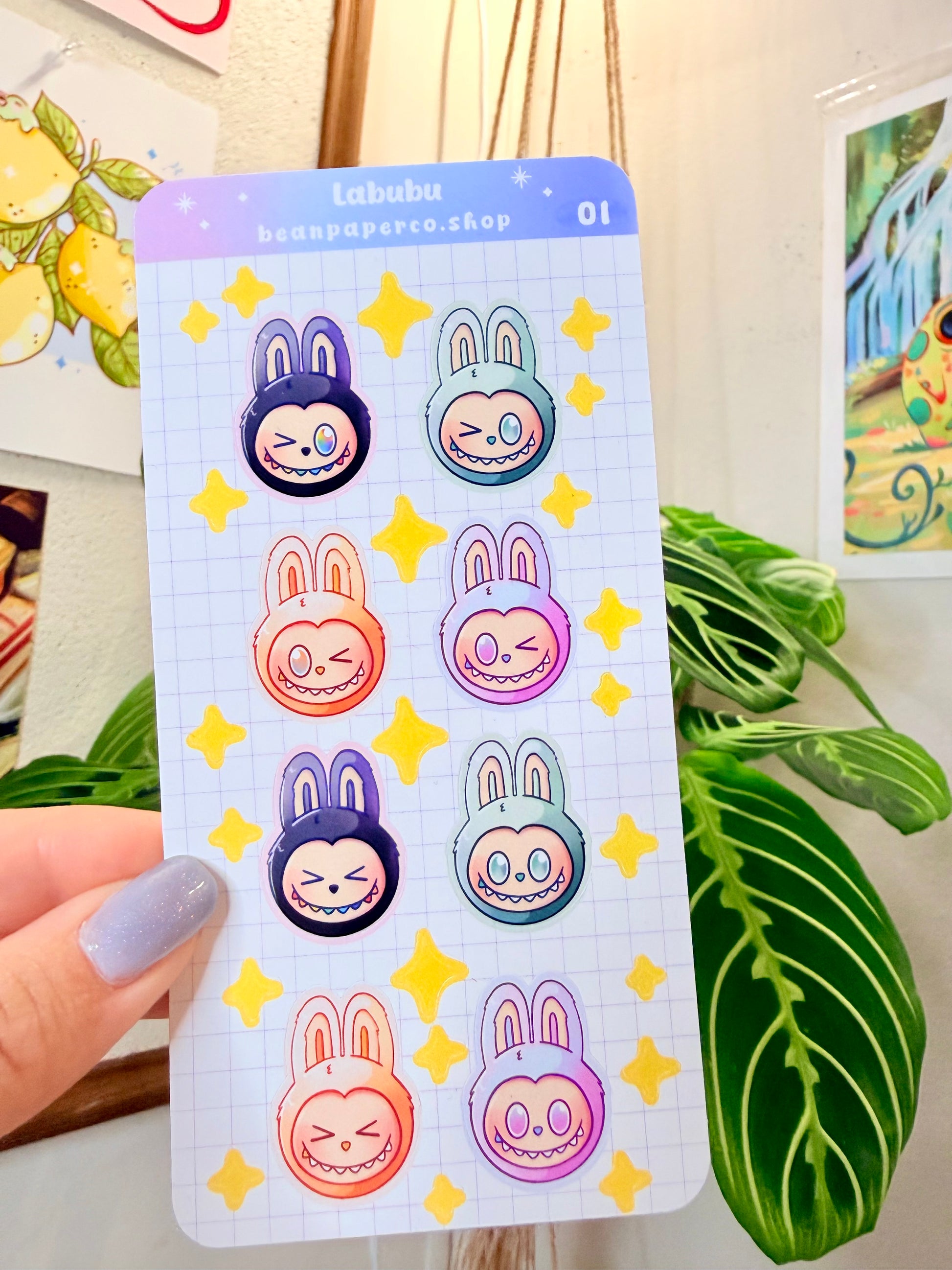 Labubu The Monster Sticker Sheet with black, blue green, orange, and purple labubus held by hand with plant in the background.