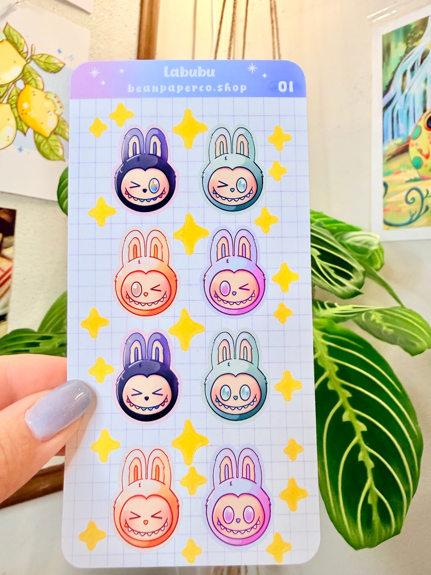 Labubu The Monster Sticker Sheet with black, blue green, orange, and purple labubus held by hand with plant in the background.
