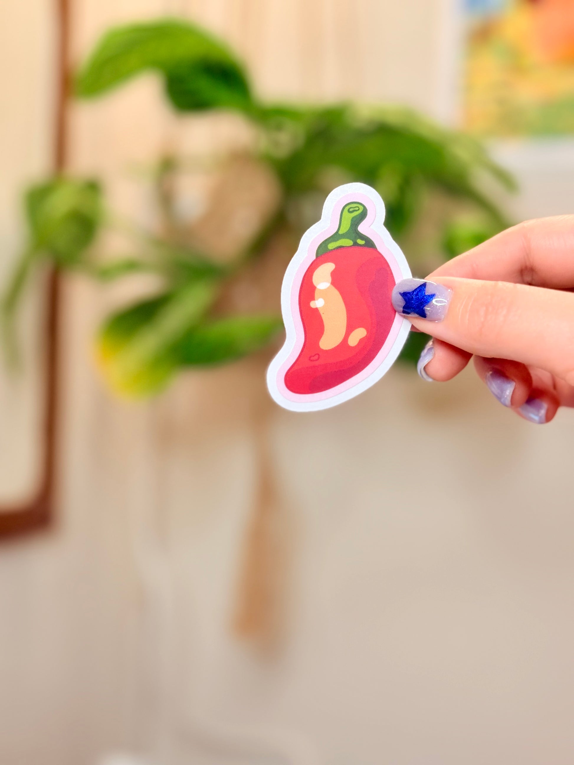 Hand holding a sticker of a chili pepper with pink outline against a blurred indoor background