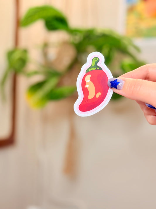 Hand holding a sticker of a chili pepper with pink outline against a blurred indoor background