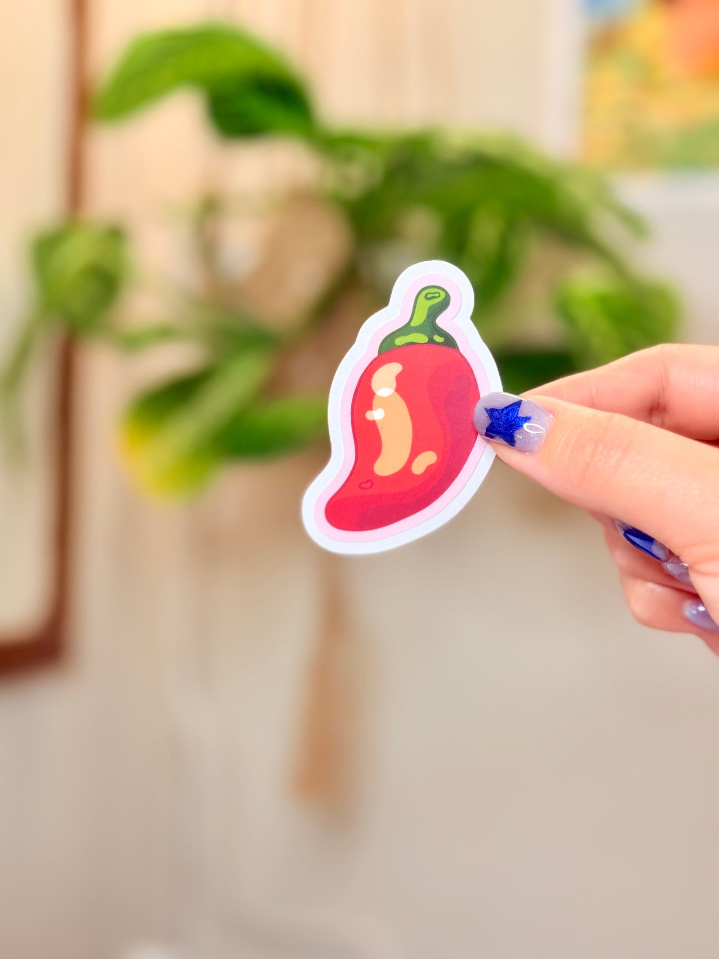 Hand holding a sticker of a chili pepper with pink outline against a blurred indoor background