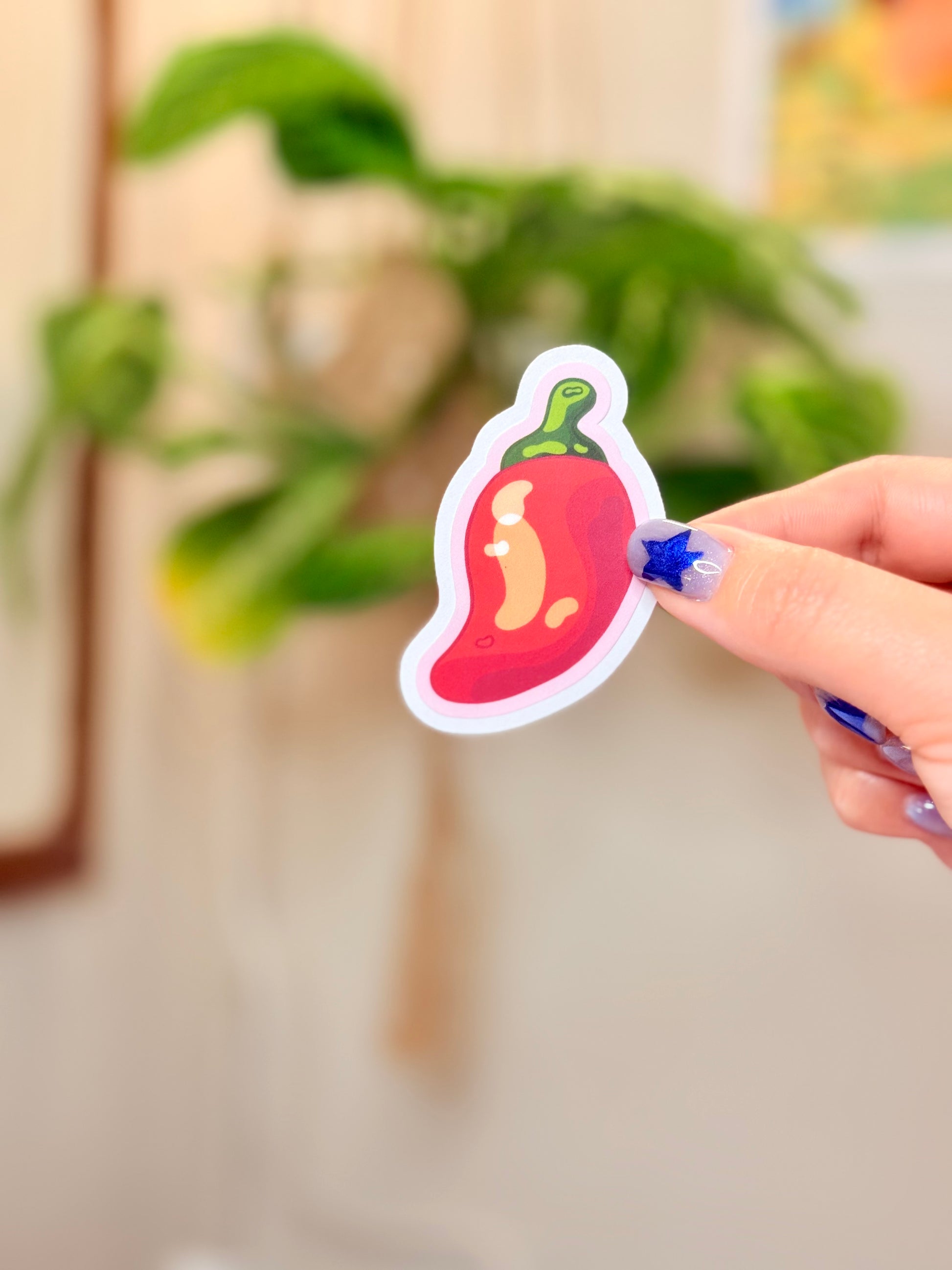 Hand holding a sticker of a chili pepper with pink outline against a blurred indoor background