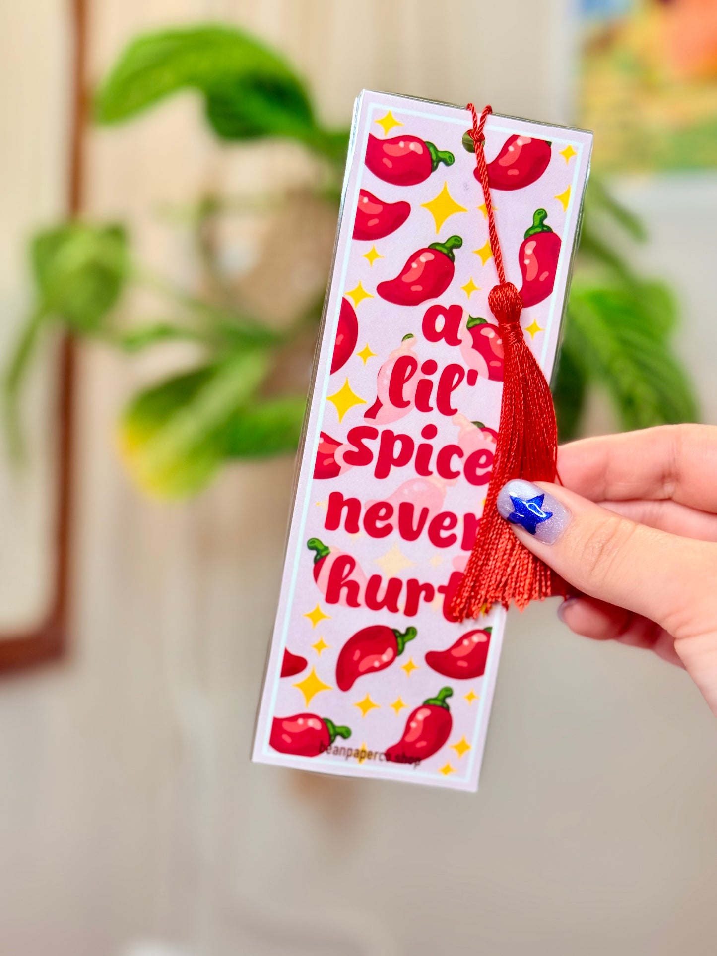 Bookmark with red peppers, a red tassel and text that says "a lil spice never hurt," held by a hand, with a plant in the background