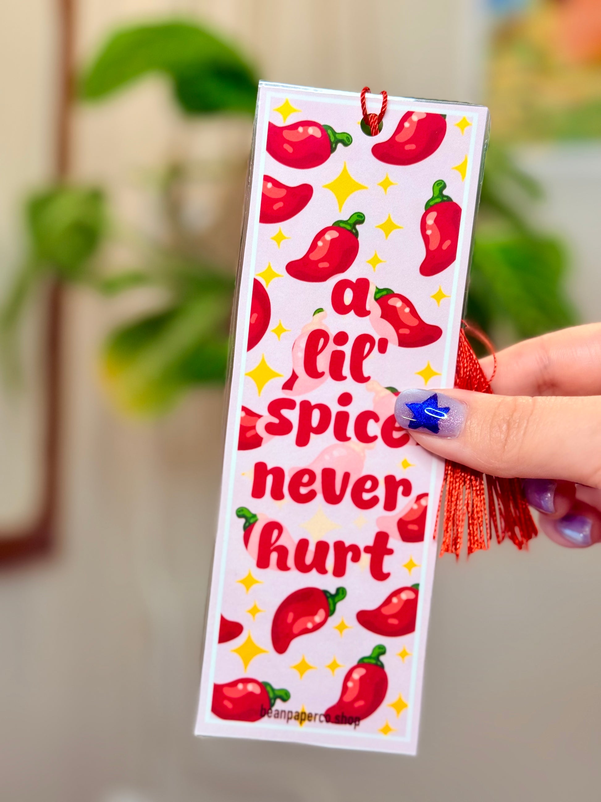 Bookmark with red peppers, a red tassel and text that says "a lil spice never hurt," held by a hand, with a plant in the background