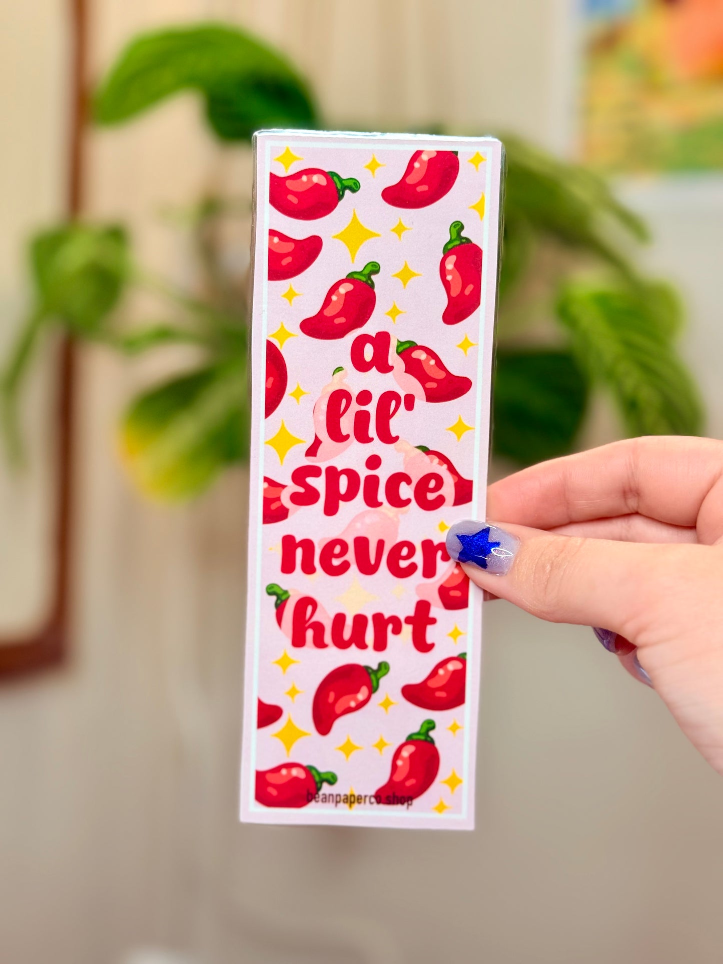 Bookmark with red peppers and text that says "a lil spice never hurt," held by a hand, with a plant in the background