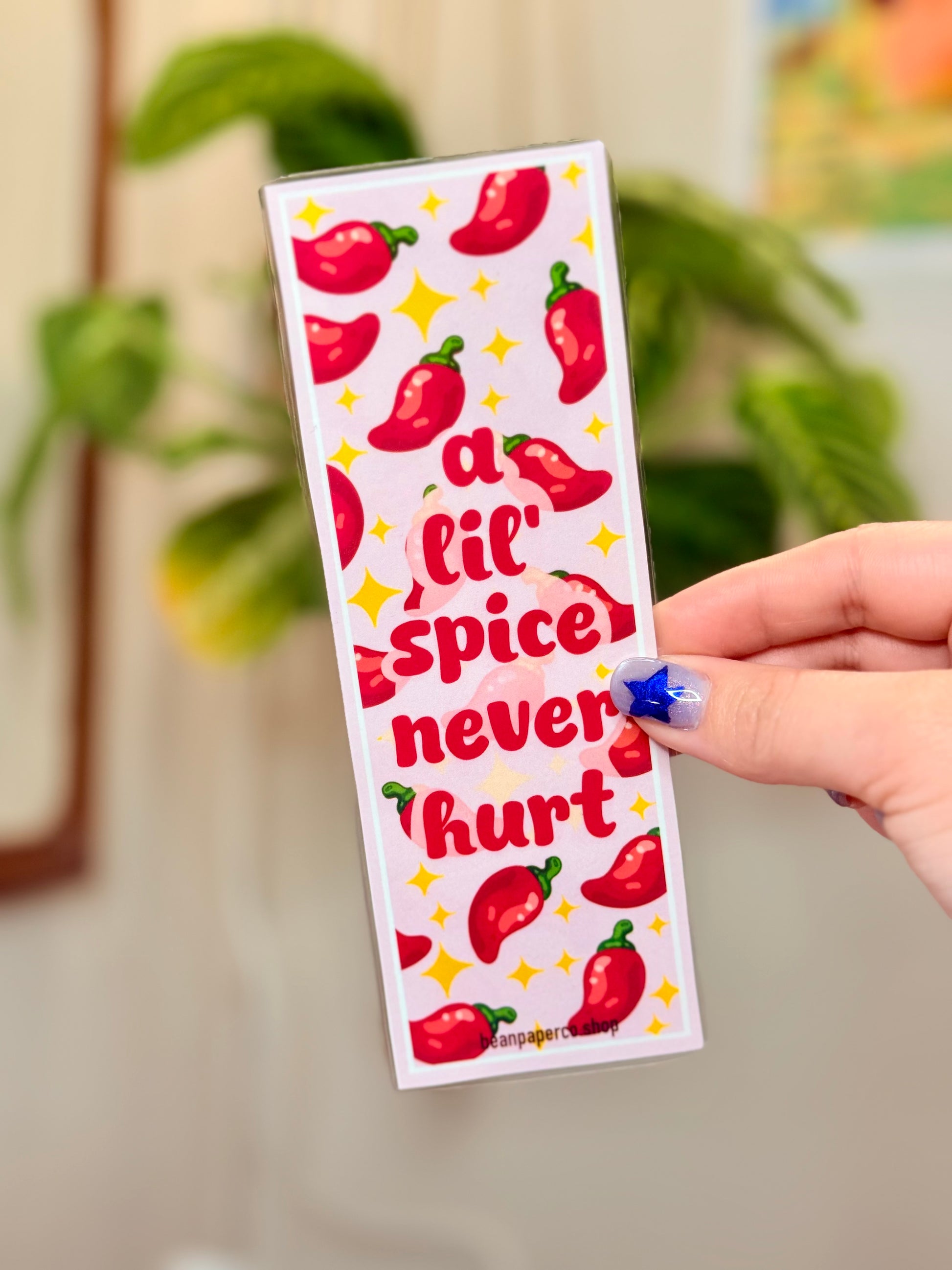 Bookmark with red peppers and text that says "a lil spice never hurt," held by a hand, with a plant in the background