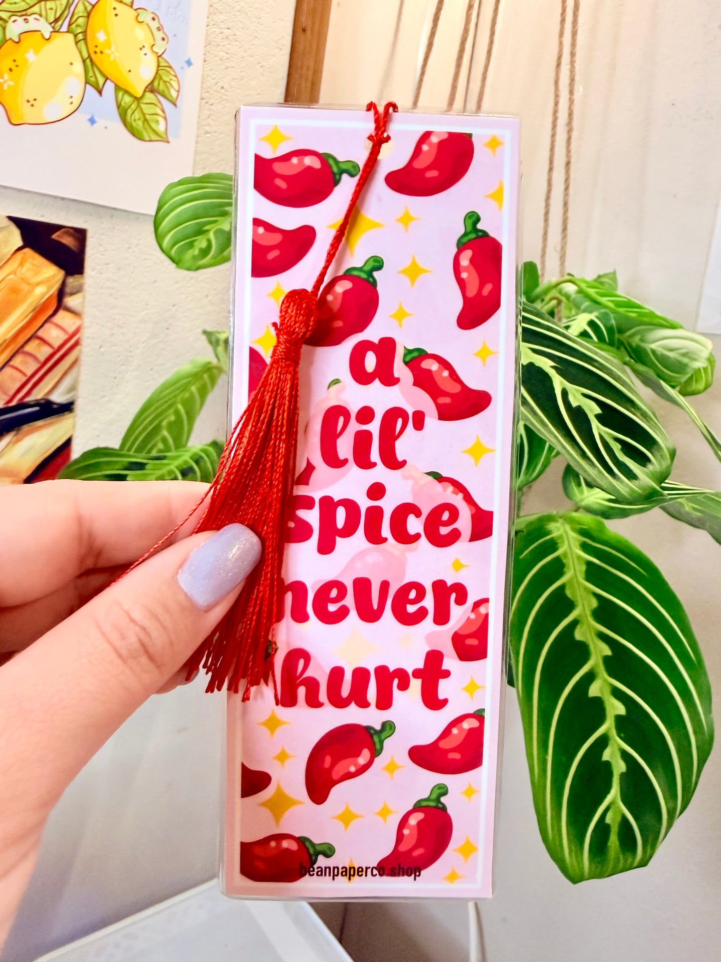 Bookmark with red peppers, a red tassel and text that says "a lil spice never hurt," held by a hand, with a plant in the background