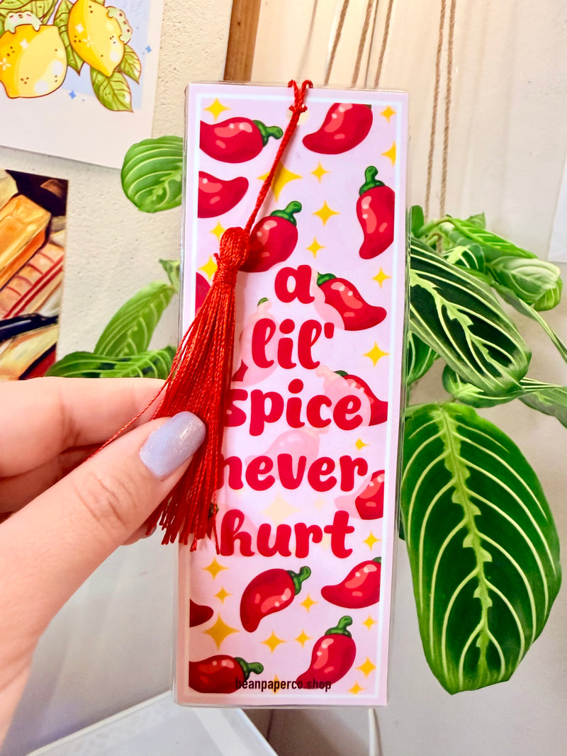 Bookmark with red peppers, a red tassel and text that says "a lil spice never hurt," held by a hand, with a plant in the background