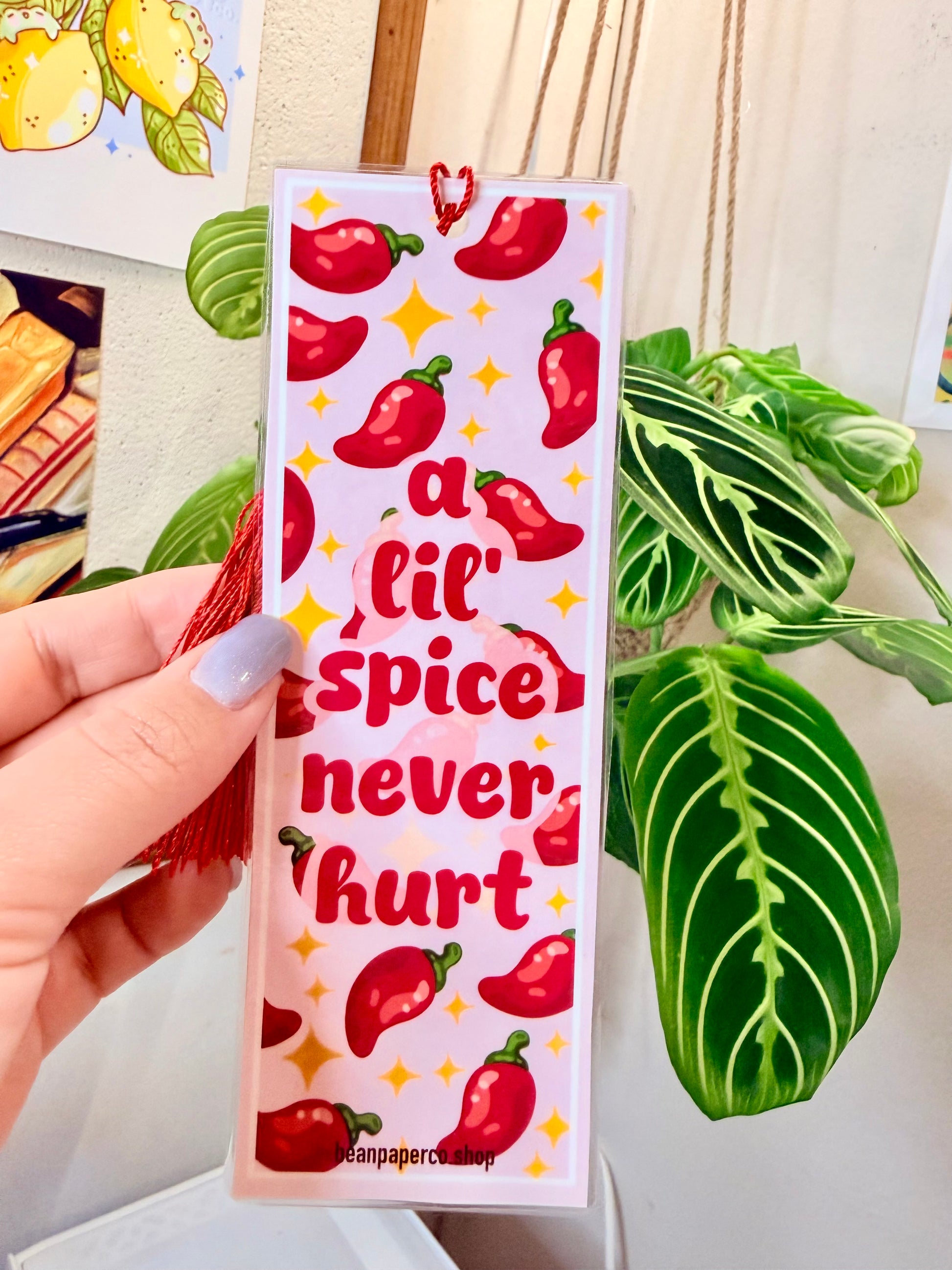 Bookmark with red peppers, a red tassel and text that says "a lil spice never hurt," held by a hand, with a plant in the background