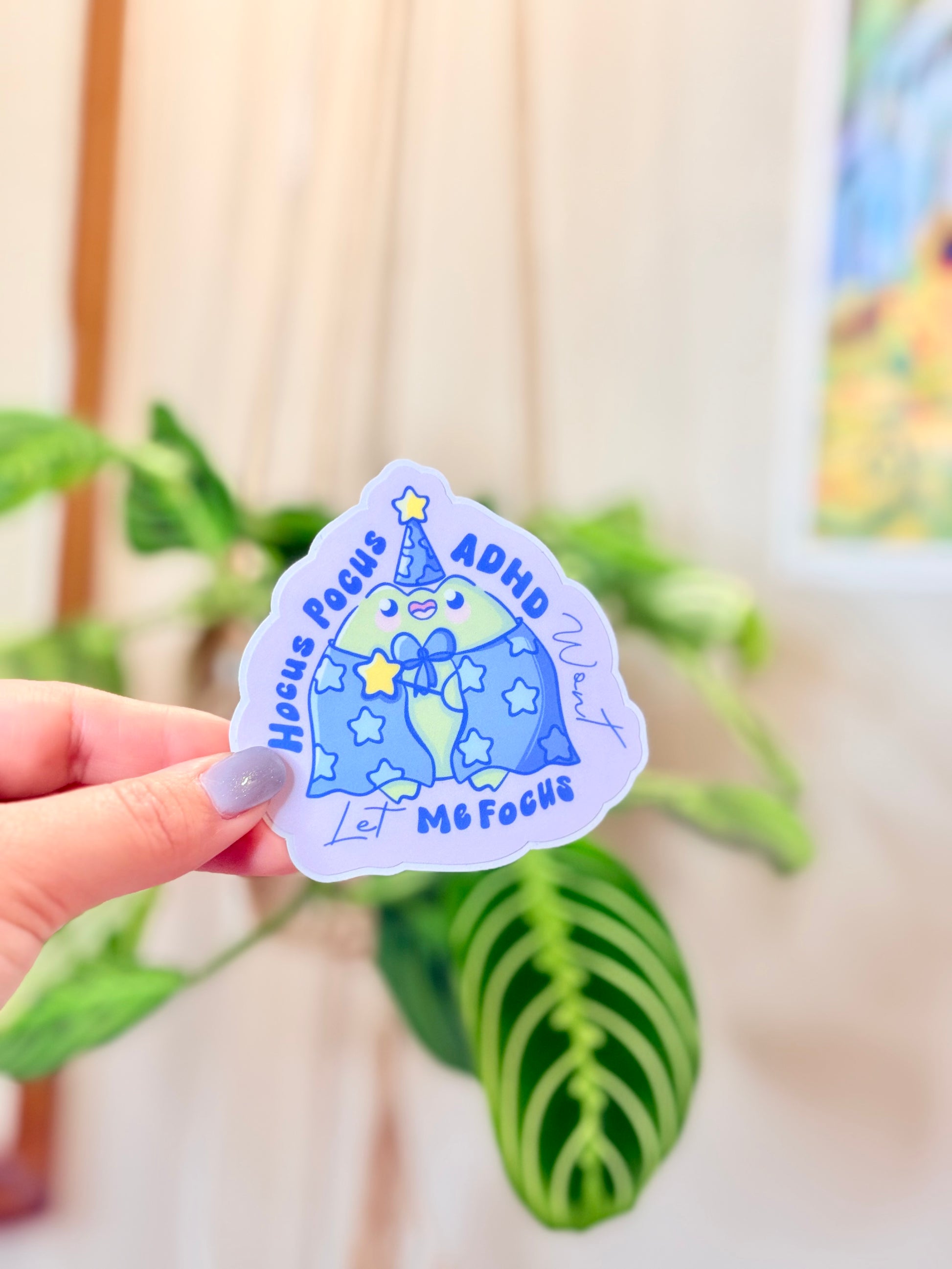 Cute ADHD Focus Frog Sticker Cute Neurodivergent Vinyl Sticker held in hand with green plants in the background.