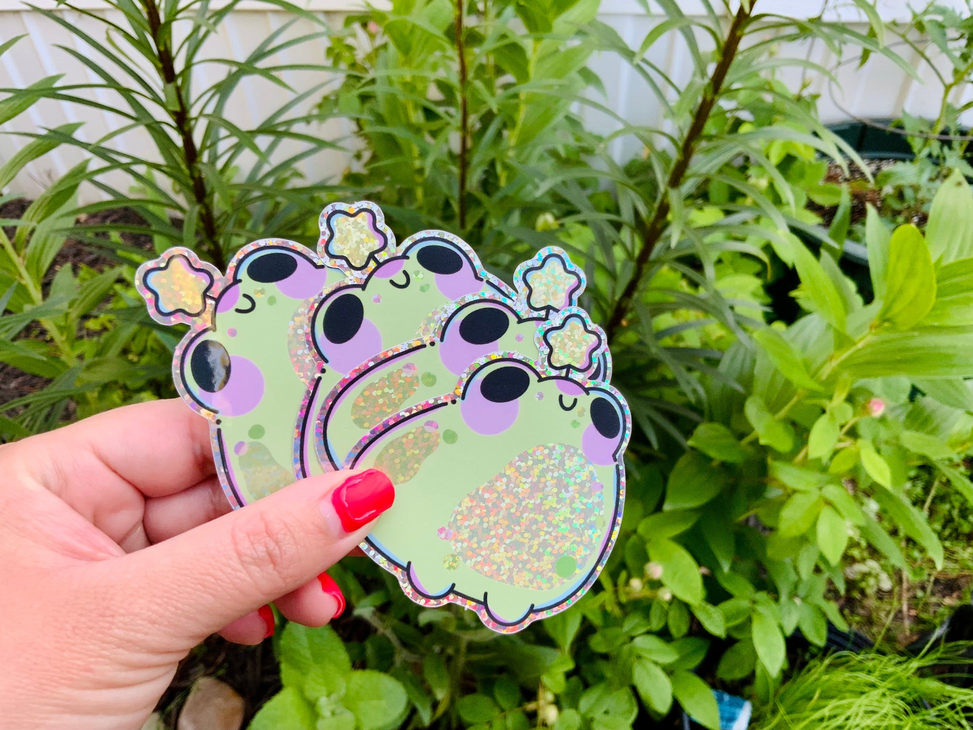 Hand holding a glittery green frog sticker with purple cheeks and a sparkly star detail, photographed outdoors against leafy green plants.