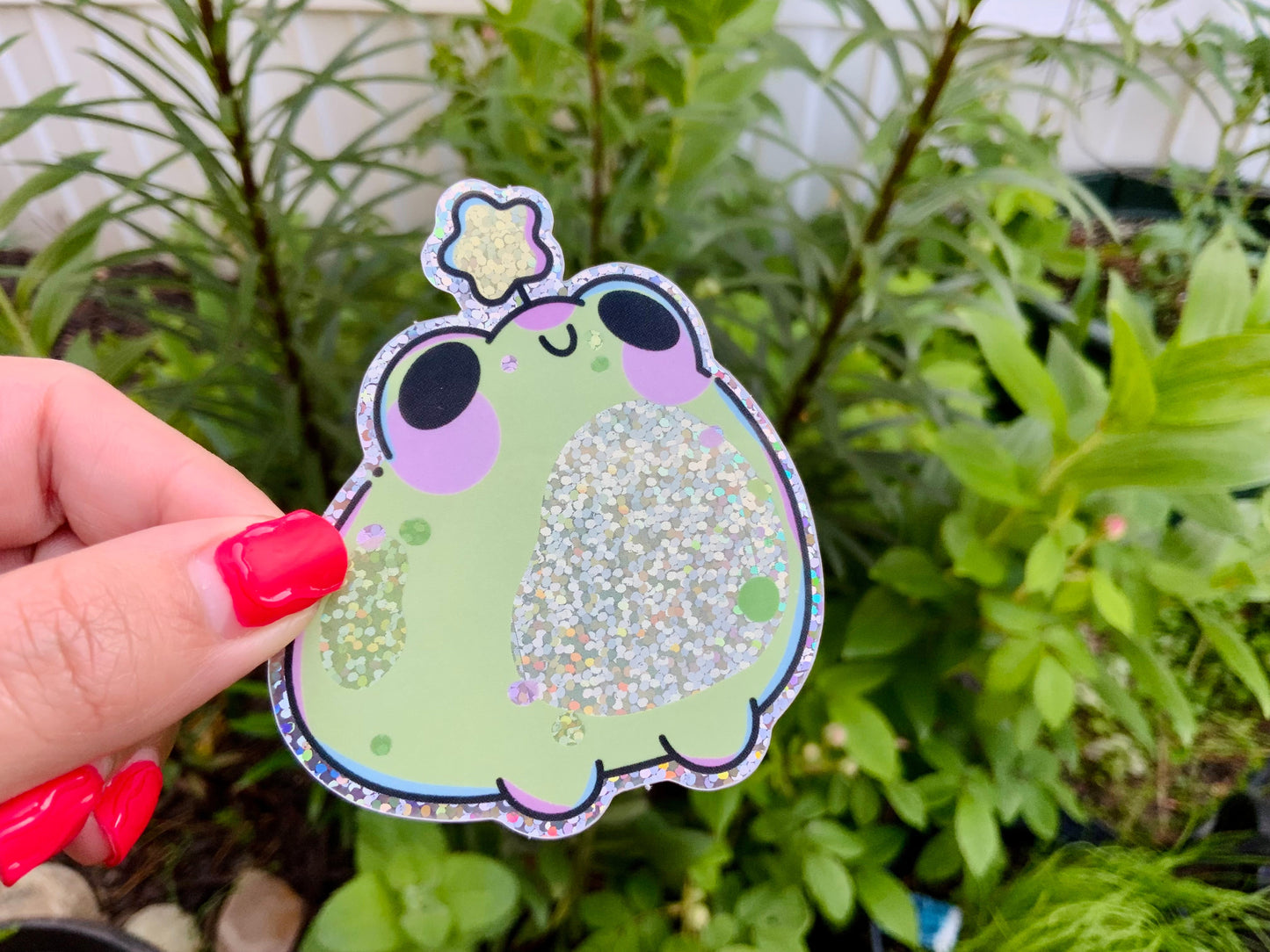 Hand holding a glittery green frog sticker with purple cheeks and a sparkly star detail, photographed outdoors against leafy green plants.