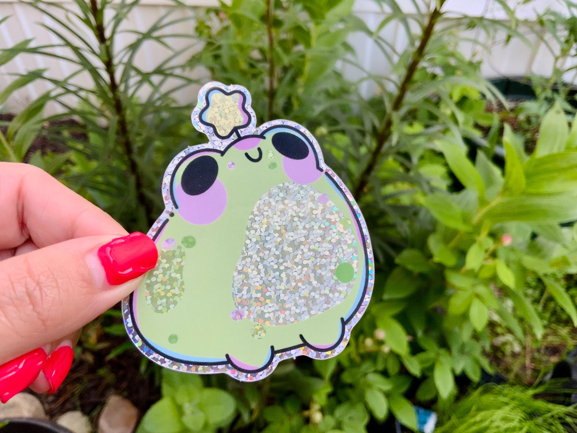 Hand holding a glittery green frog sticker with purple cheeks and a sparkly star detail, photographed outdoors against leafy green plants.