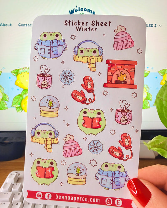 “Winter frog sticker sheet featuring frogs in scarves and earmuffs, mittens, snow globes, tea mugs, hats, snowflakes, and a cozy fireplace.”