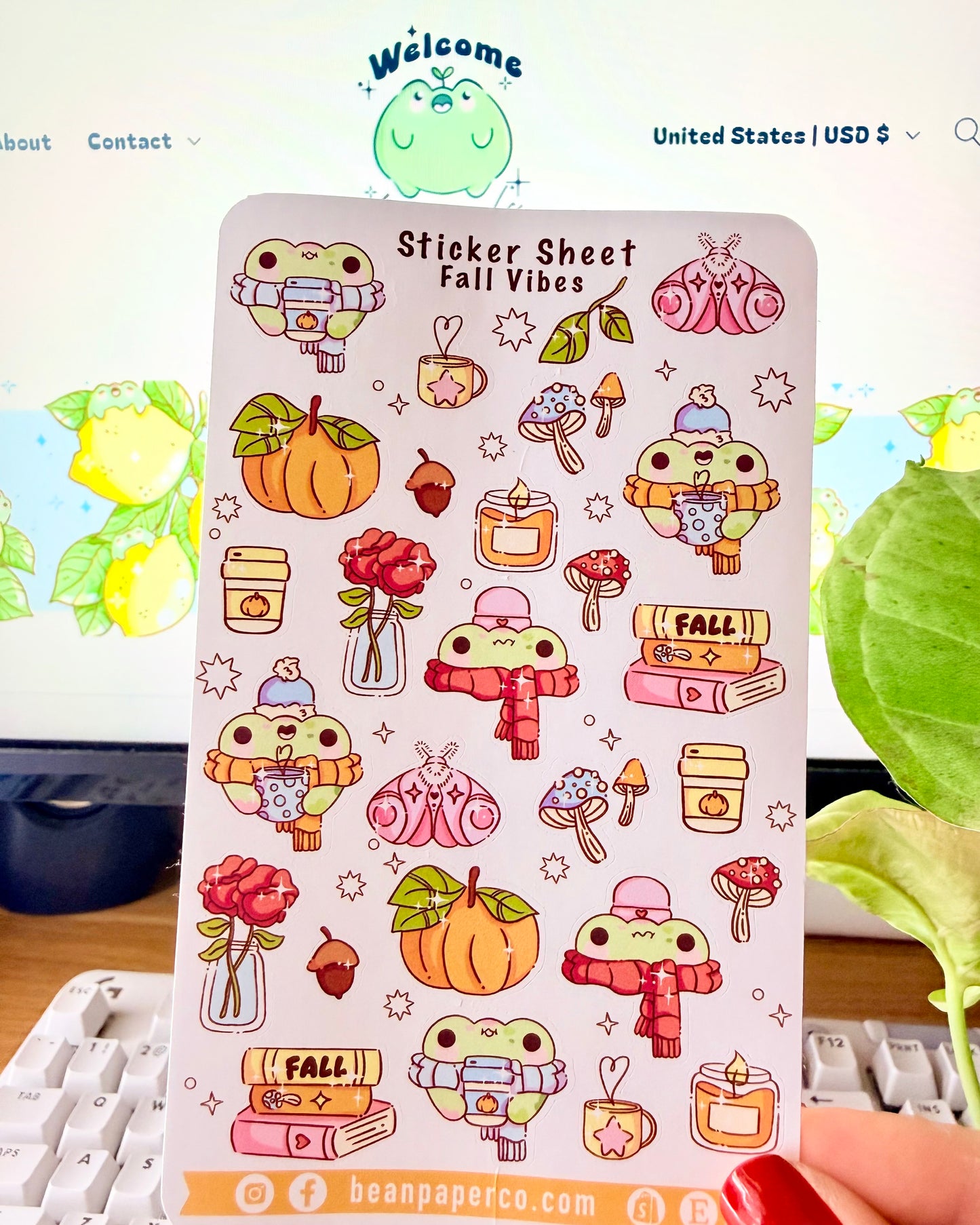 Sticker sheet titled “Sticker Sheet Fall Vibes” featuring cute frog illustrations with pumpkins, scarves, mushrooms, candles, books, acorns, roses, and cozy autumn drinks, held in front of a desk setup.