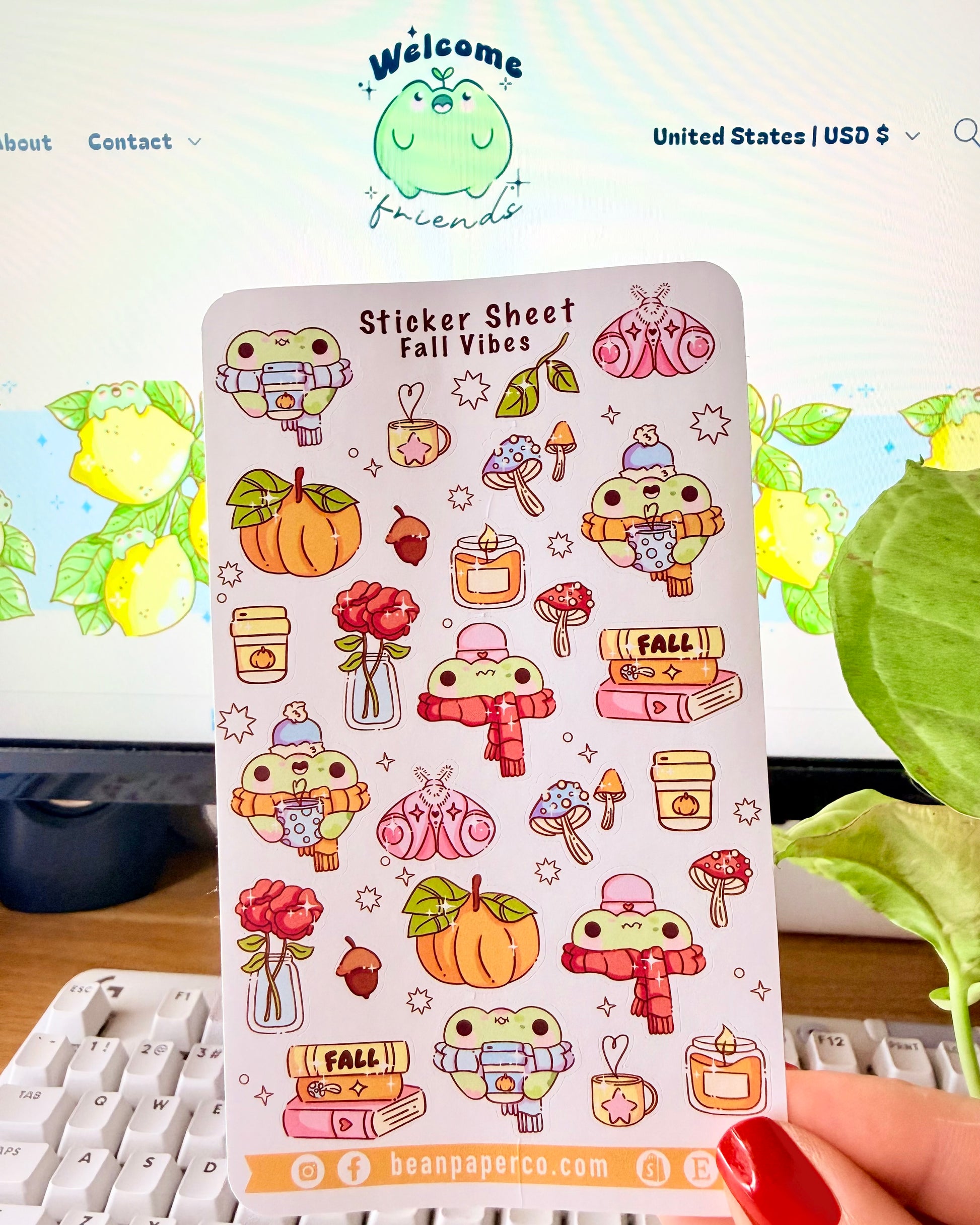 Sticker sheet titled “Sticker Sheet Fall Vibes” featuring cute frog illustrations with pumpkins, scarves, mushrooms, candles, books, acorns, roses, and cozy autumn drinks, held in front of a desk setup.