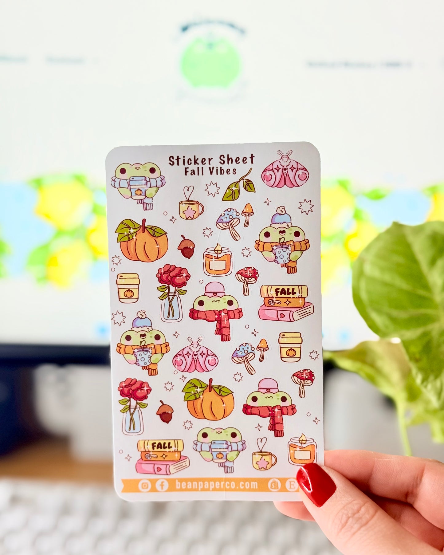 Sticker sheet titled “Sticker Sheet Fall Vibes” featuring cute frog illustrations with pumpkins, scarves, mushrooms, candles, books, acorns, roses, and cozy autumn drinks, held in front of a desk setup.
