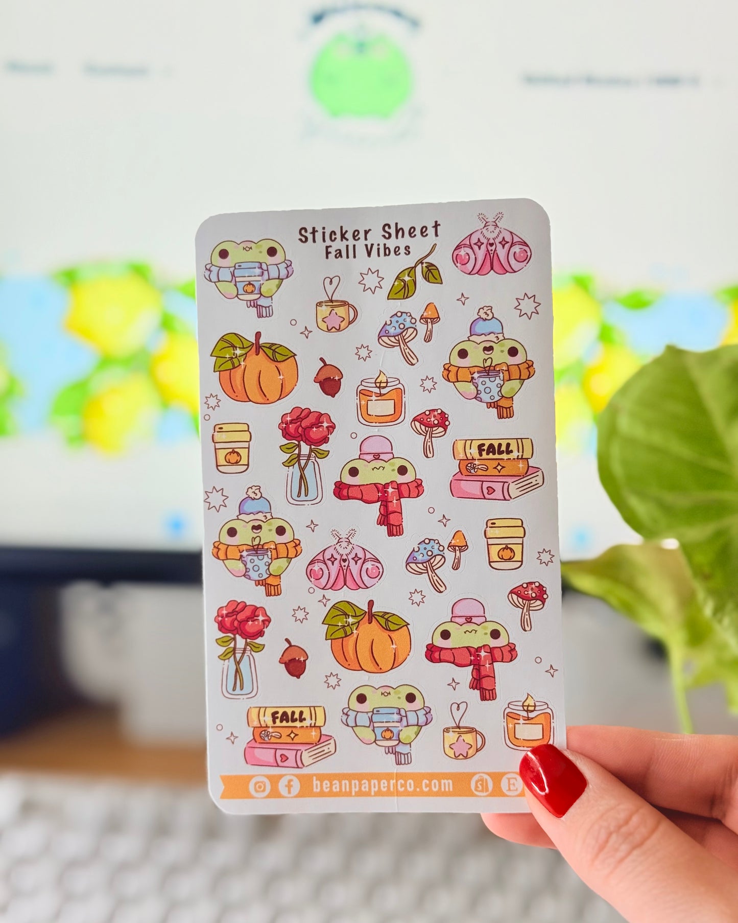 Sticker sheet titled “Sticker Sheet Fall Vibes” featuring cute frog illustrations with pumpkins, scarves, mushrooms, candles, books, acorns, roses, and cozy autumn drinks, held in front of a desk setup.