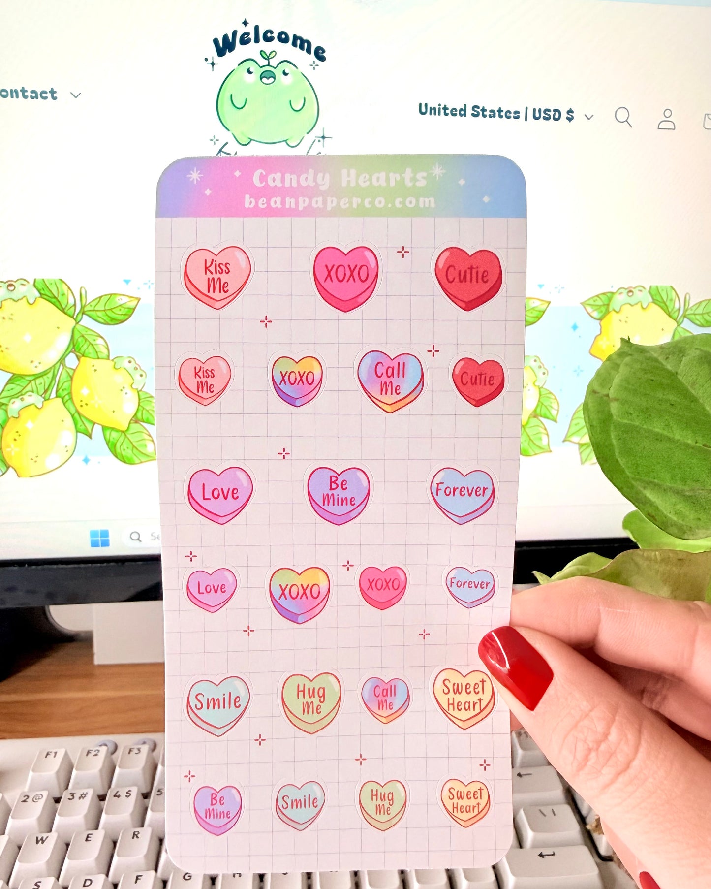 Valentine Heart Candy Sticker Sheet. Hand holding a Valentine’s candy heart sticker sheet featuring pastel conversation hearts with phrases like “Kiss Me,” “XOXO,” “Be Mine,” and “Sweet Heart,” photographed in front of a computer screen and plant.