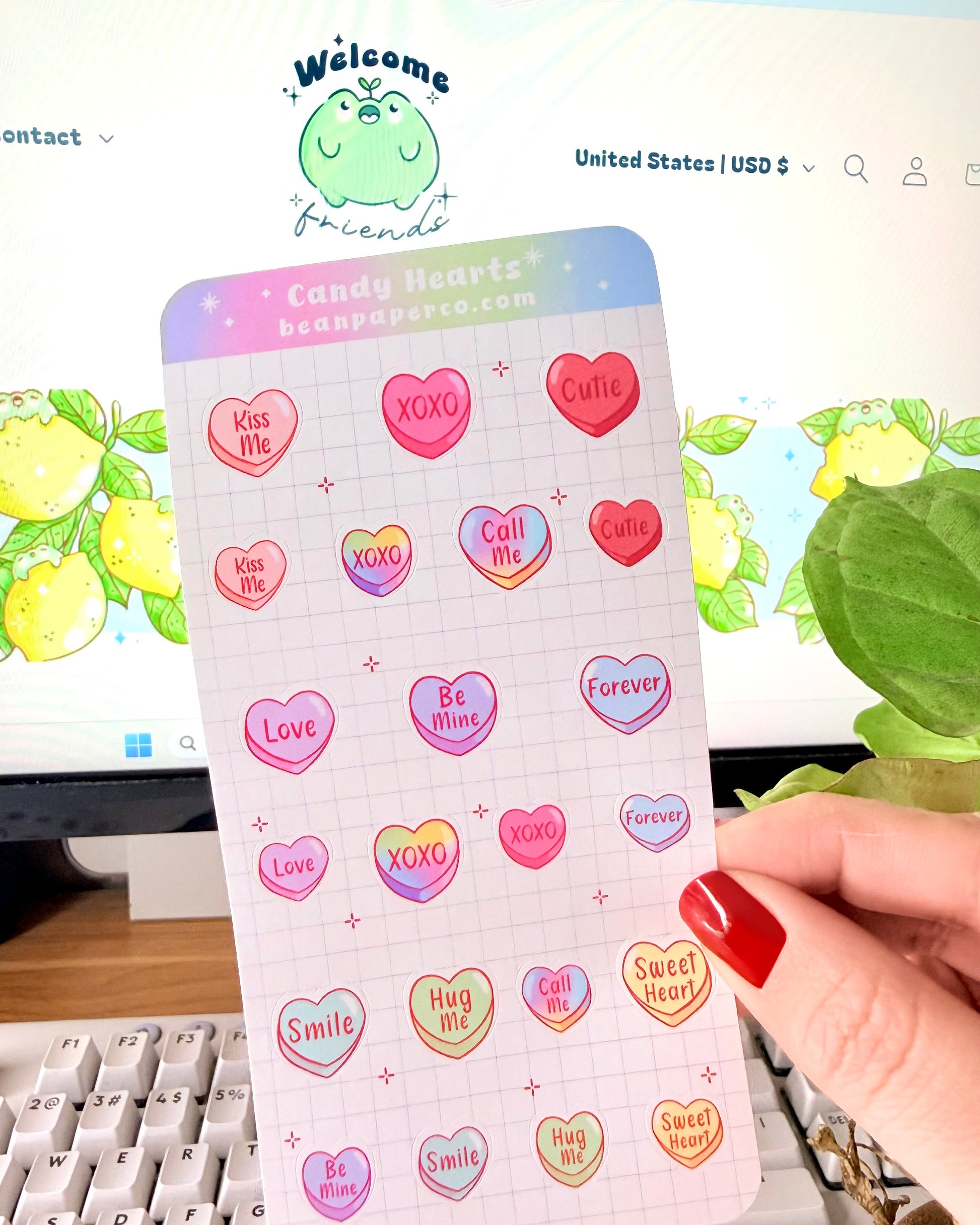 Valentine Heart Candy Sticker Sheet. Hand holding a Valentine’s candy heart sticker sheet featuring pastel conversation hearts with phrases like “Kiss Me,” “XOXO,” “Be Mine,” and “Sweet Heart,” photographed in front of a computer screen and plant.