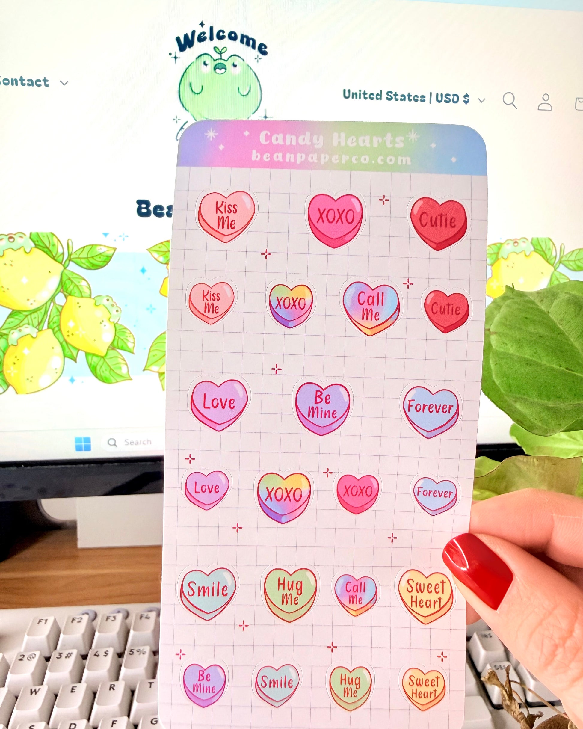 Valentine Heart Candy Sticker Sheet. Hand holding a Valentine’s candy heart sticker sheet featuring pastel conversation hearts with phrases like “Kiss Me,” “XOXO,” “Be Mine,” and “Sweet Heart,” photographed in front of a computer screen and plant.