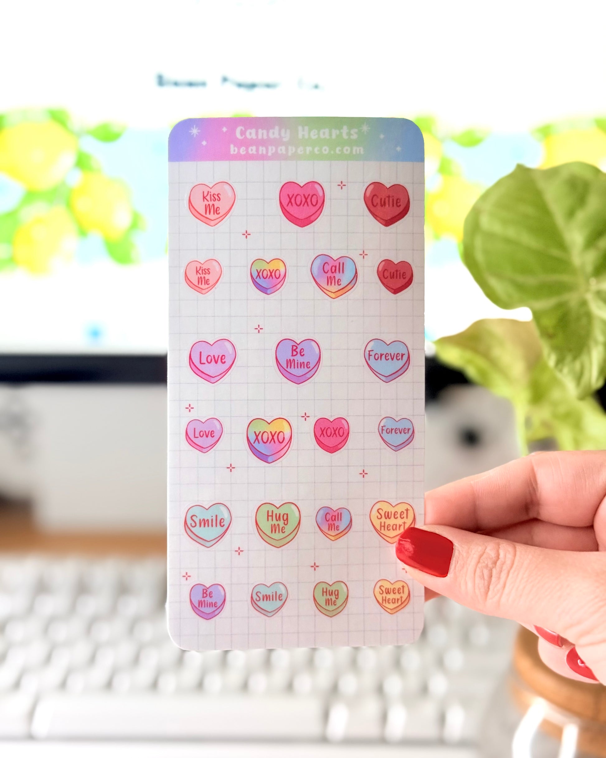 Valentine Heart Candy Sticker Sheet. Hand holding a Valentine’s candy heart sticker sheet featuring pastel conversation hearts with phrases like “Kiss Me,” “XOXO,” “Be Mine,” and “Sweet Heart,” photographed in front of a computer screen and plant.