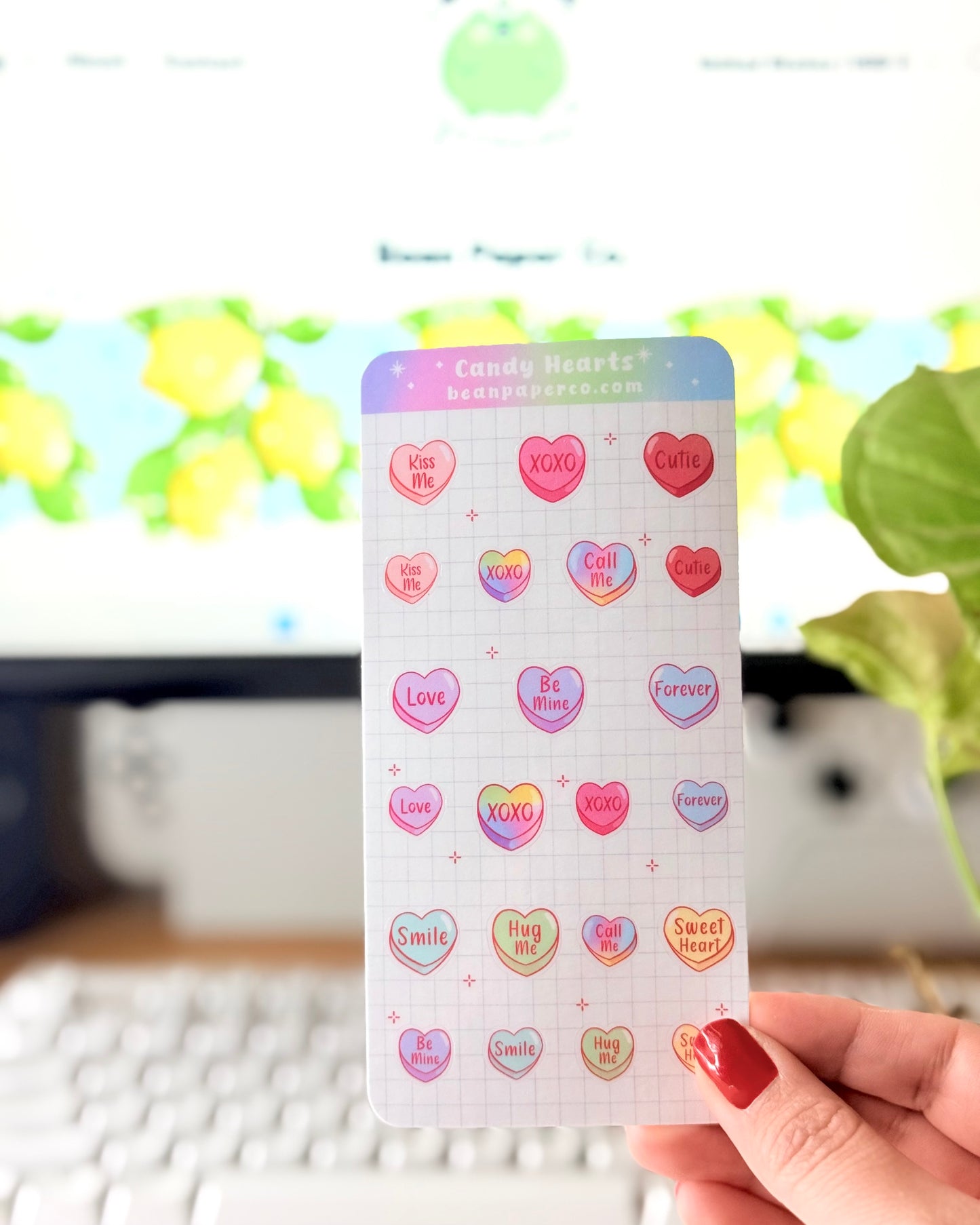Valentine Heart Candy Sticker Sheet. Hand holding a Valentine’s candy heart sticker sheet featuring pastel conversation hearts with phrases like “Kiss Me,” “XOXO,” “Be Mine,” and “Sweet Heart,” photographed in front of a computer screen and plant.