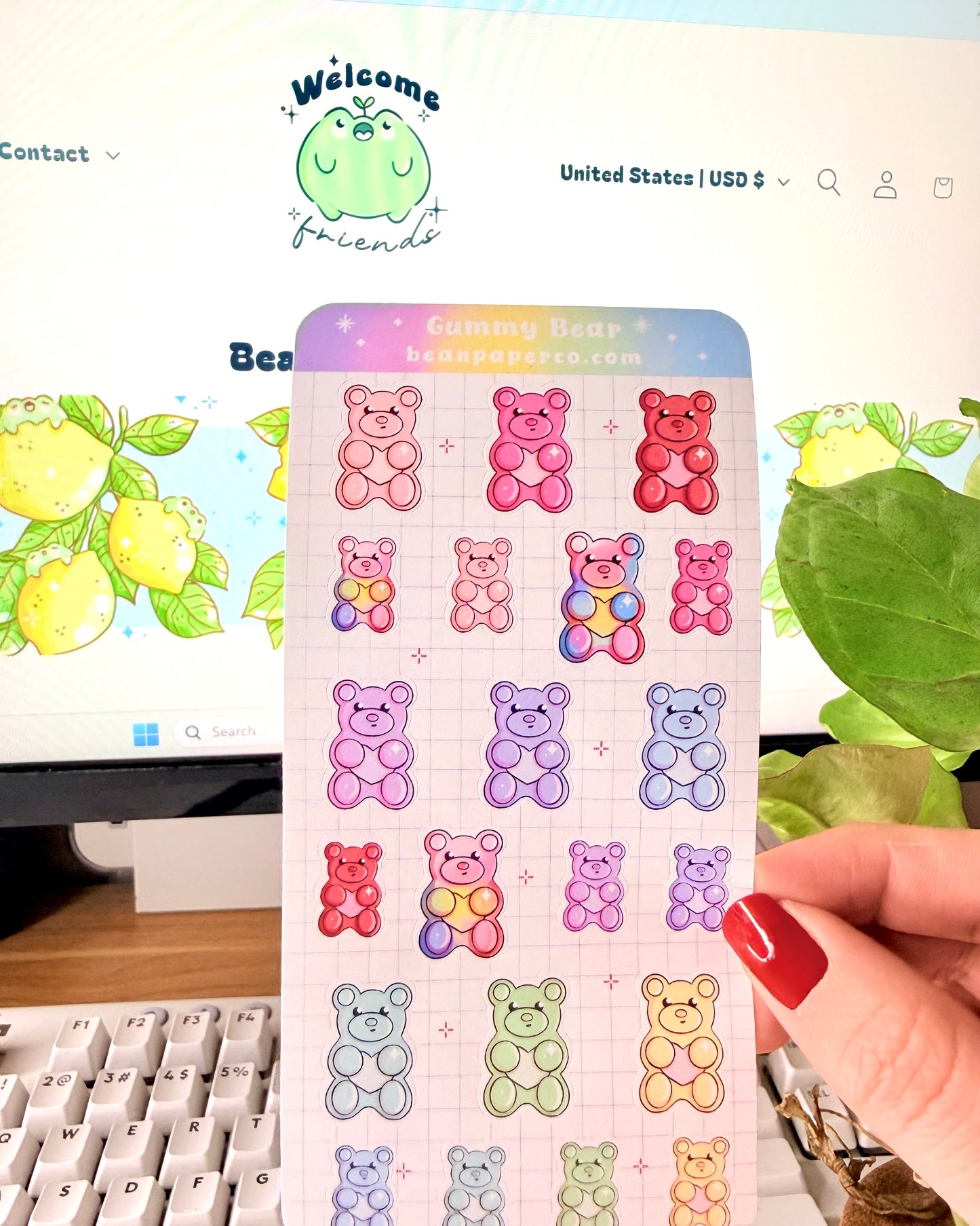 Valentine Gummy Bear Sticker Sheet. Hand holding a pastel gummy bear sticker sheet with multiple colorful bear designs in pink, purple, blue, green, and red on a grid background, photographed in front of a computer screen and plant.