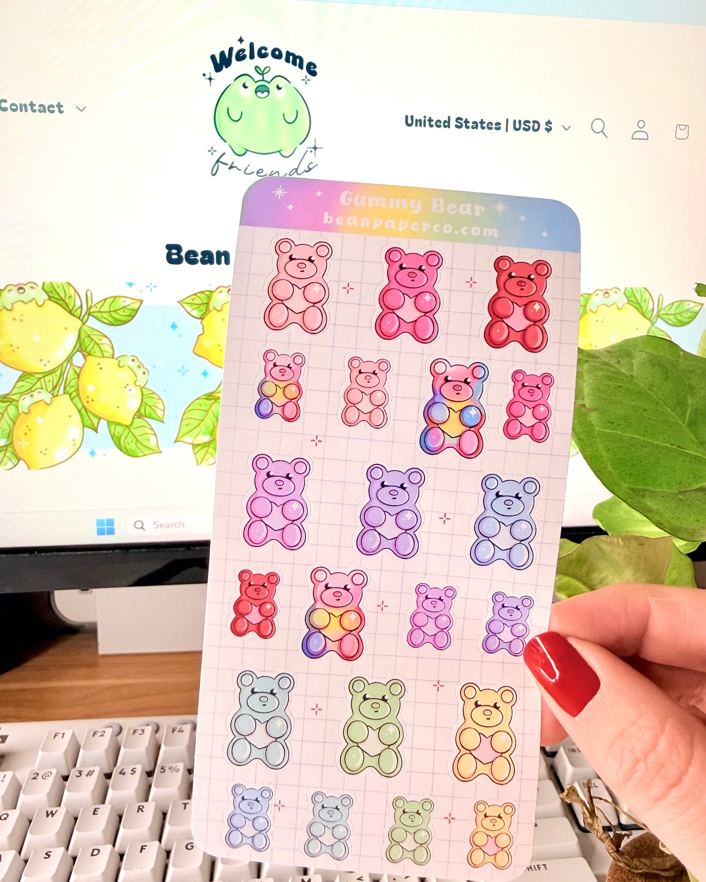 Valentine Gummy Bear Sticker Sheet. Hand holding a pastel gummy bear sticker sheet with multiple colorful bear designs in pink, purple, blue, green, and red on a grid background, photographed in front of a computer screen and plant.