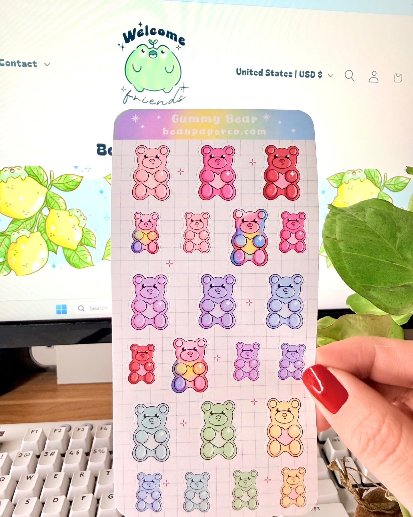Valentine Gummy Bear Sticker Sheet. Hand holding a pastel gummy bear sticker sheet with multiple colorful bear designs in pink, purple, blue, green, and red on a grid background, photographed in front of a computer screen and plant.