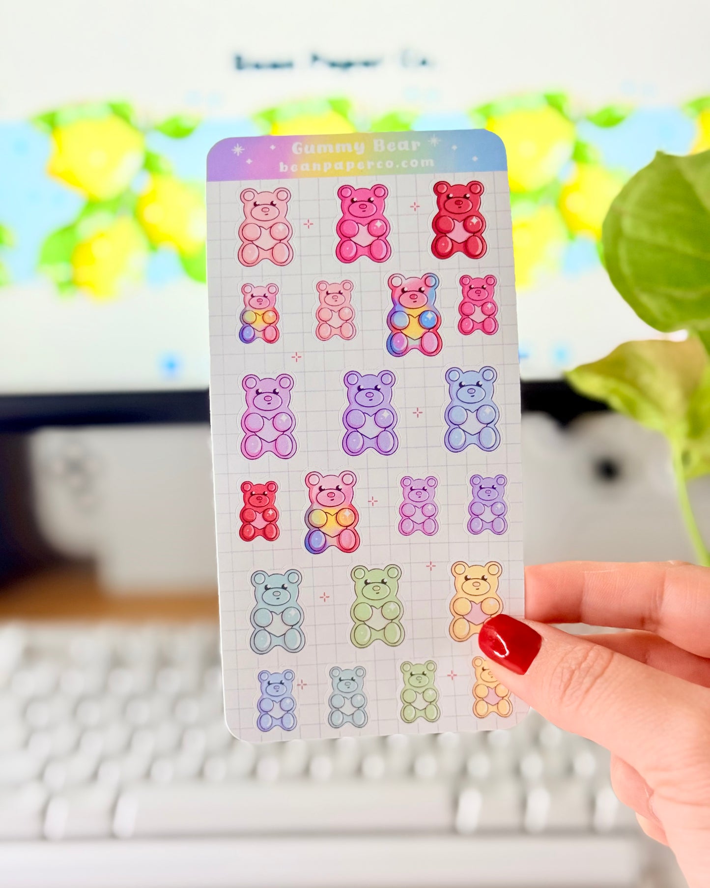 Valentine Gummy Bear Sticker Sheet. Hand holding a pastel gummy bear sticker sheet with multiple colorful bear designs in pink, purple, blue, green, and red on a grid background, photographed in front of a computer screen and plant.