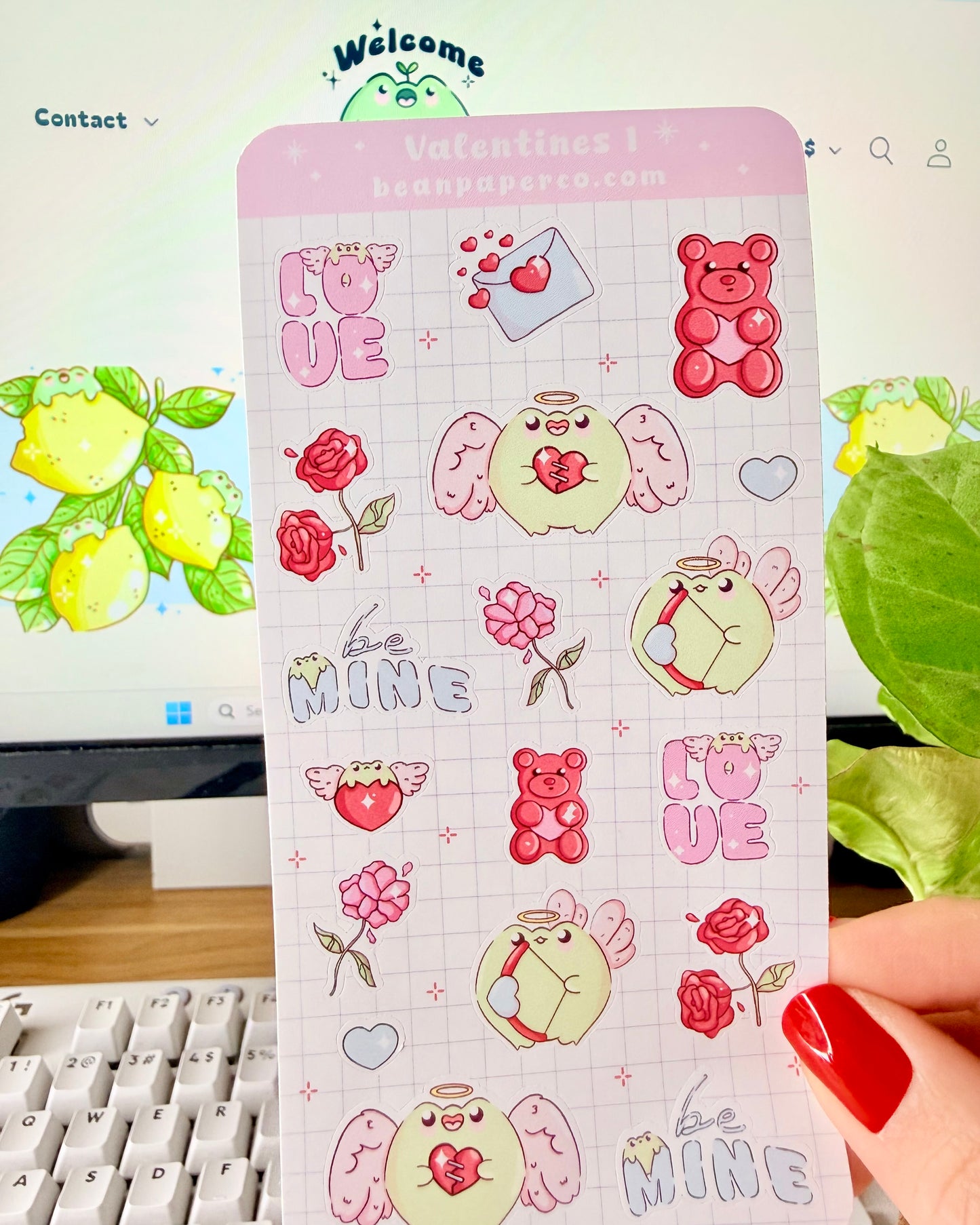Kawaii Valentine Frog Sticker Sheet. Hand holding a Valentine-themed sticker sheet featuring angel frogs, roses, gummy bears, love letters, and “Be Mine” text in pink and red tones against a computer desk background.